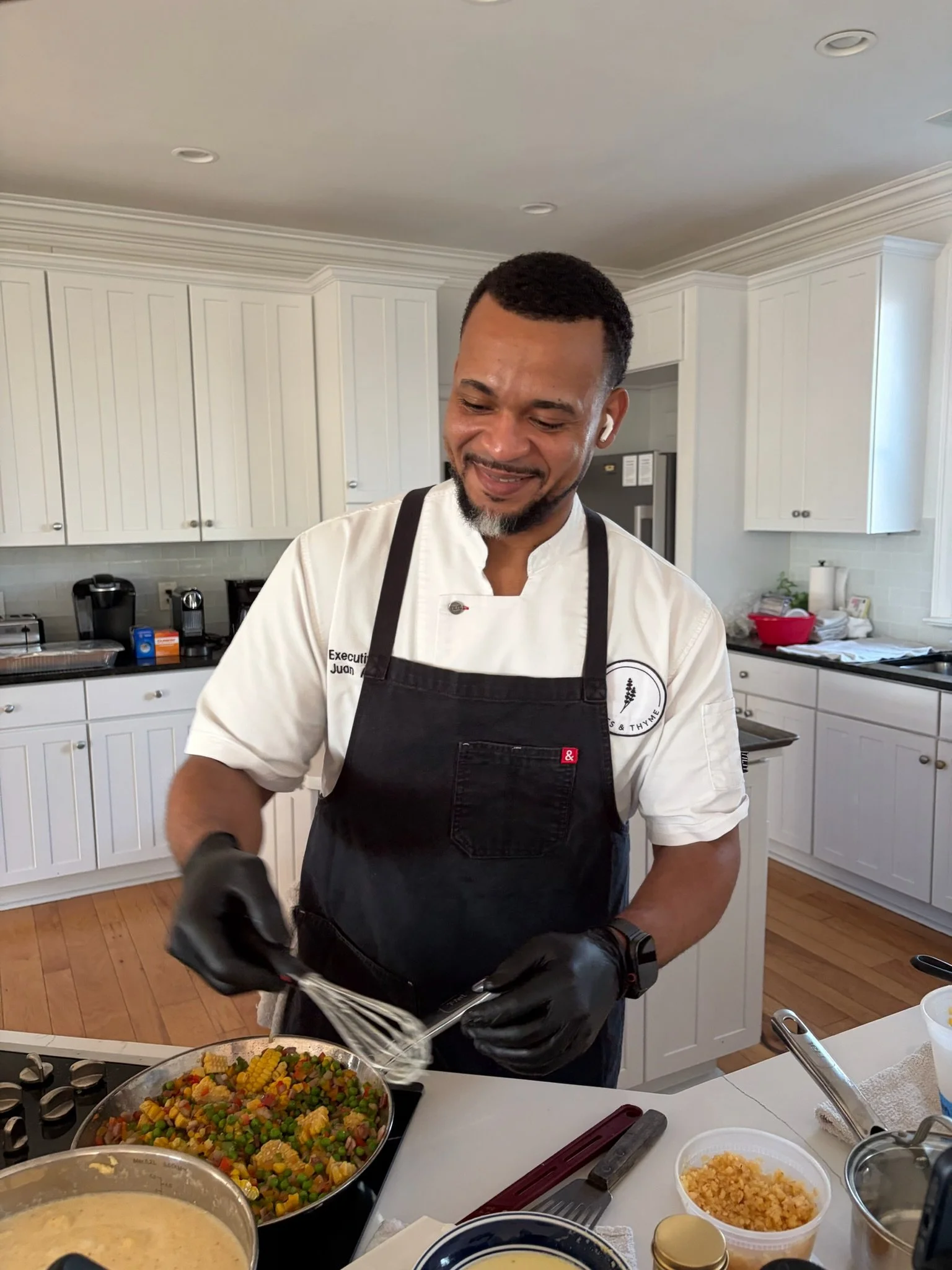 Man wearing black apron and gloves preparing food in a kitchen. On the counter are bowls with corn, rice, and mixed vegetables. The background features white cabinets and kitchen appliances.