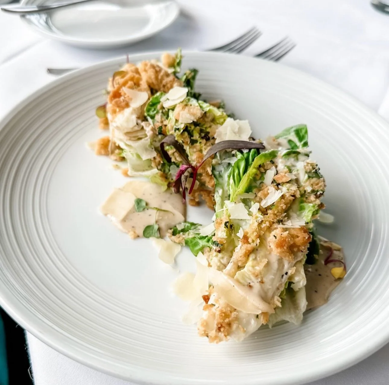 A white plate with two pieces of Caesar salad with greens, cheese, and croutons, topped with shaved cheese and a creamy dressing.