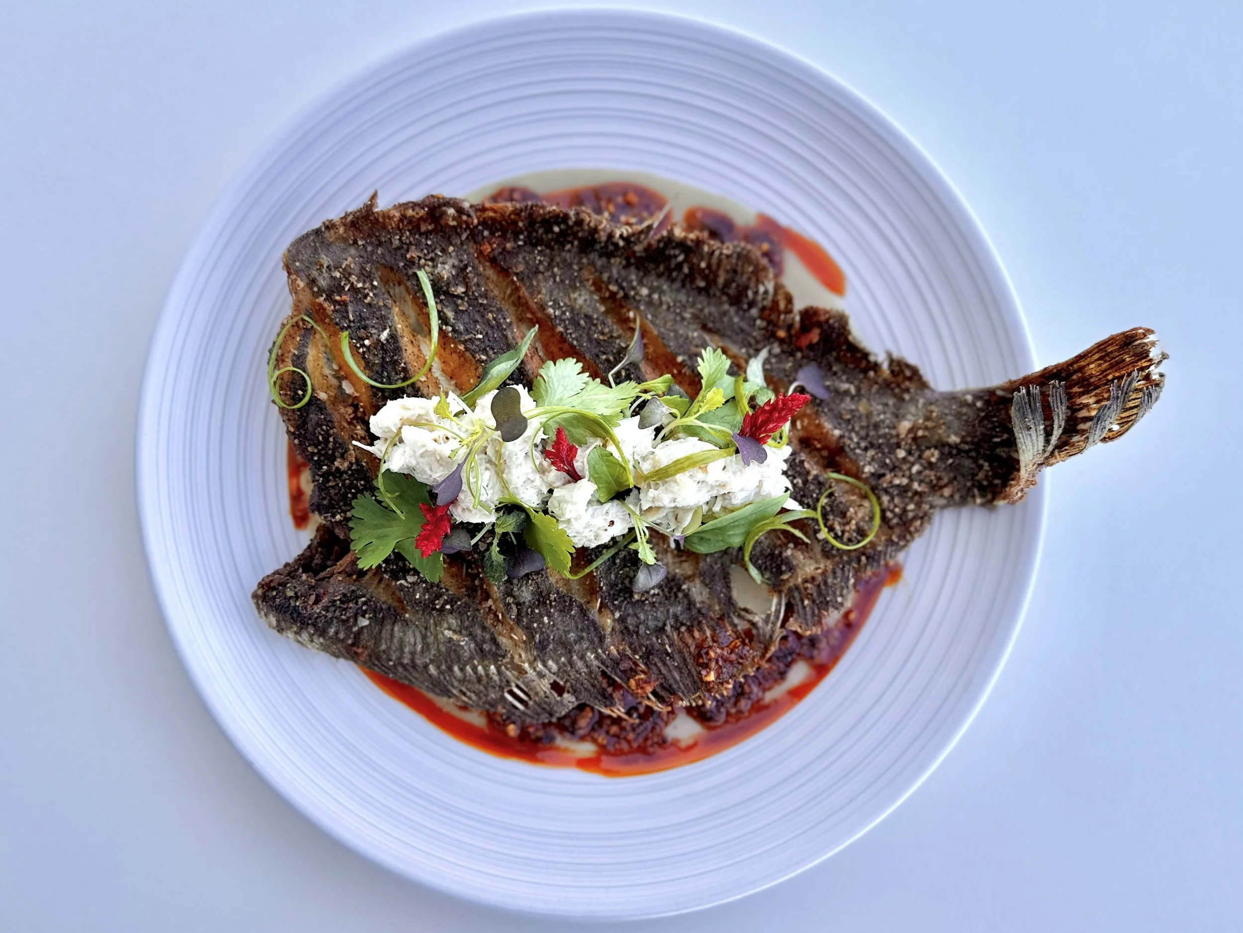 A cooked fish garnished with herbs and vegetables, served on a white plate with red sauce.