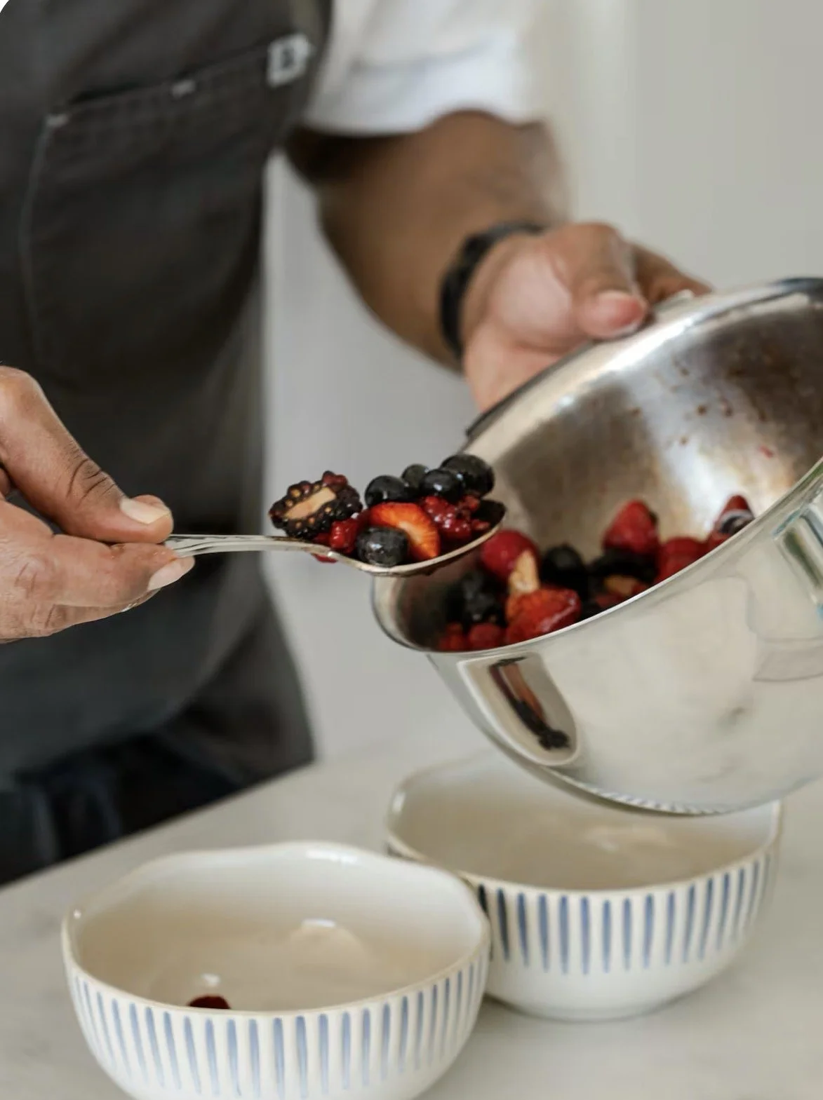 Person scooping mixed berries, including strawberries, blueberries, and blackberries, from a metal bowl into striped bowls.