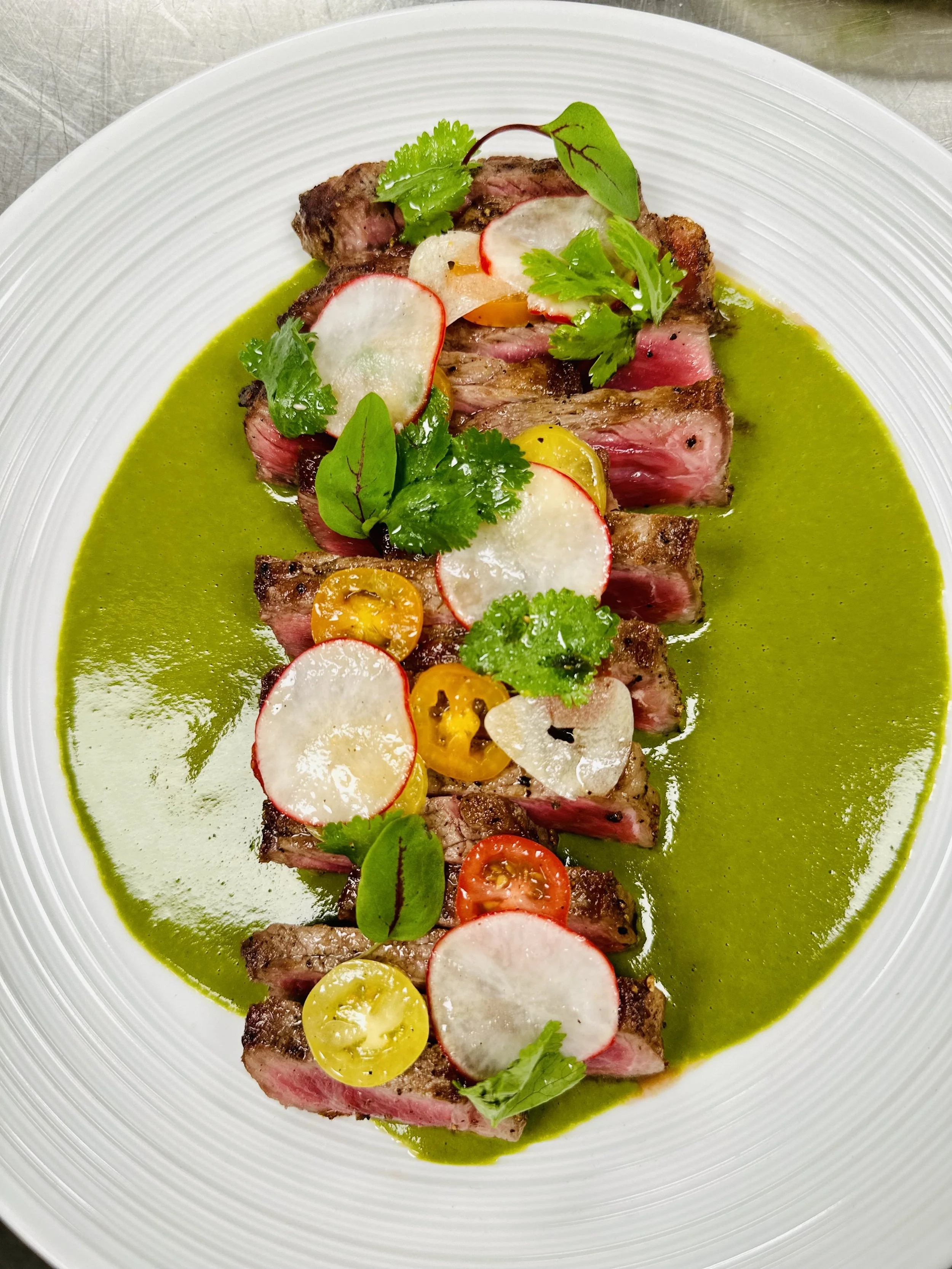 Sliced grilled steak topped with radish slices, cherry tomatoes, fresh herbs, and green sauce on a white plate.