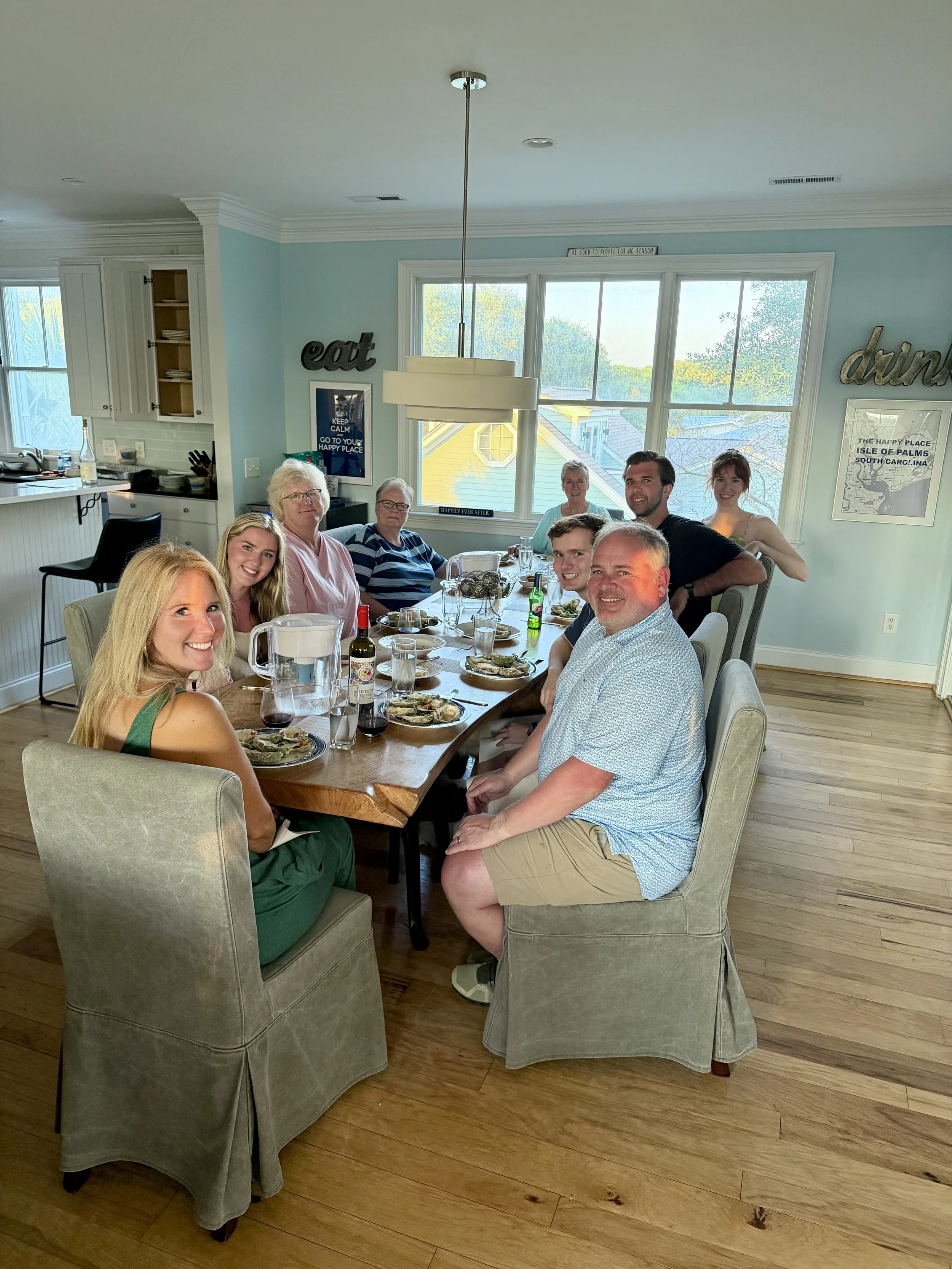 A family or group of friends gathered around a dining table in a bright, cozy kitchen and dining area, enjoying a meal together. Everyone is smiling and looking at the camera.