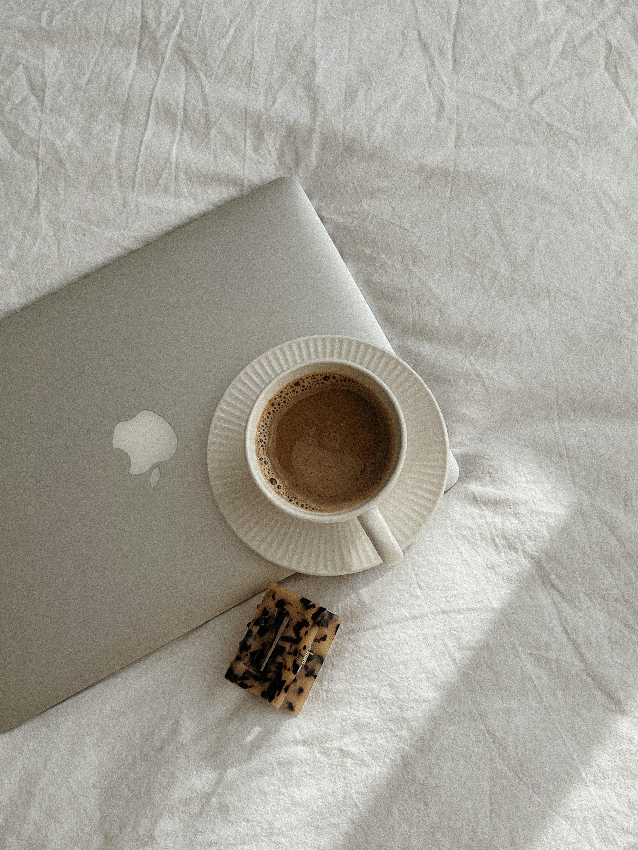 A silver laptop with an Apple logo, a white ceramic coffee cup with hot coffee on a matching saucer, a tortoise shell hair clip, all arranged on a light-colored textured fabric surface.