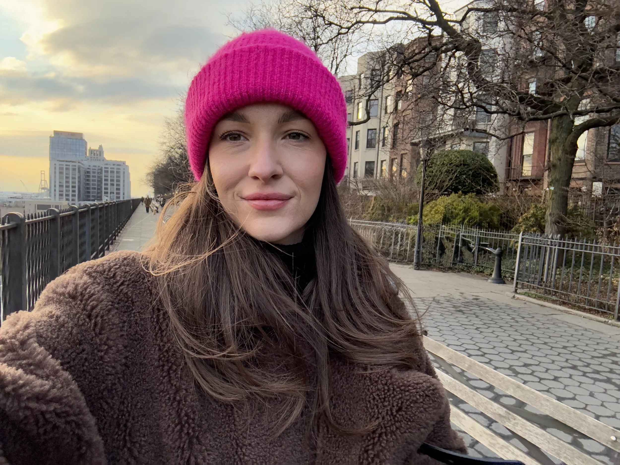 Young woman wearing a pink knit hat and a brown fuzzy coat taking a selfie on a park bench during sunset, with city buildings and trees in the background.