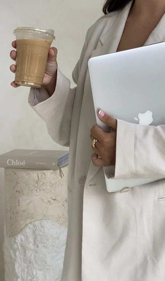 Person in a white blazer holding a plastic cup of iced coffee in one hand and a silver MacBook in the other, standing in front of a stone wall with a book titled "Chloé" on a pedestal.