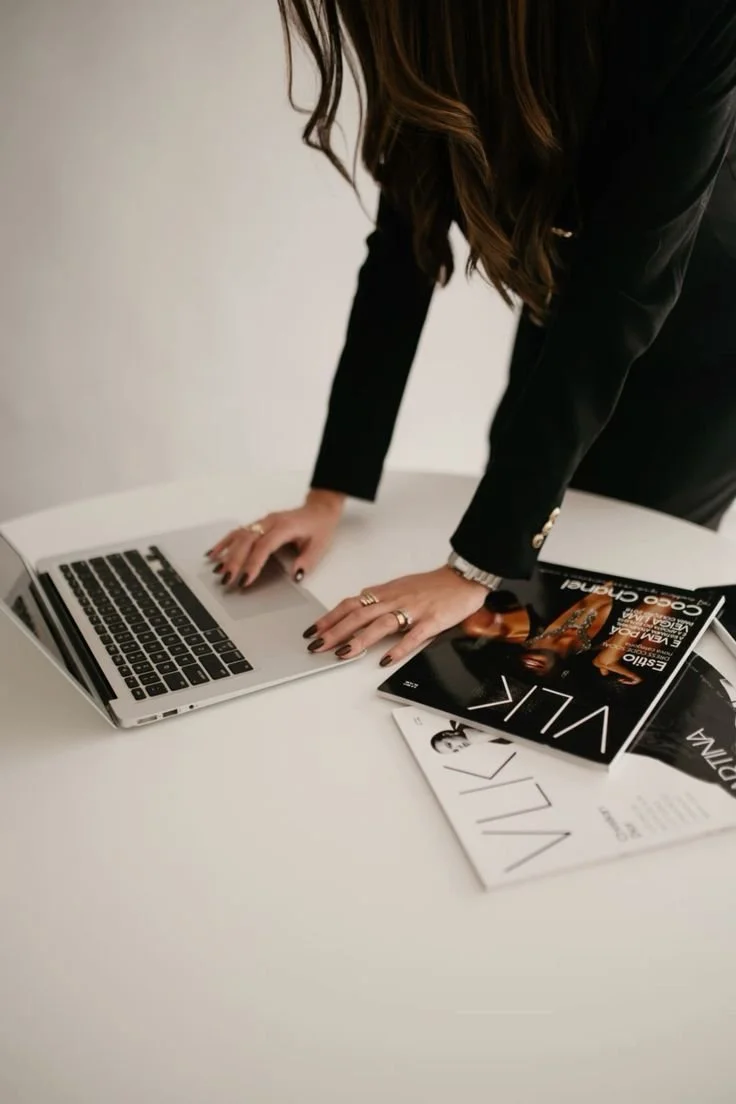 Person in black blazer leaning over white table with magazines and open laptop.