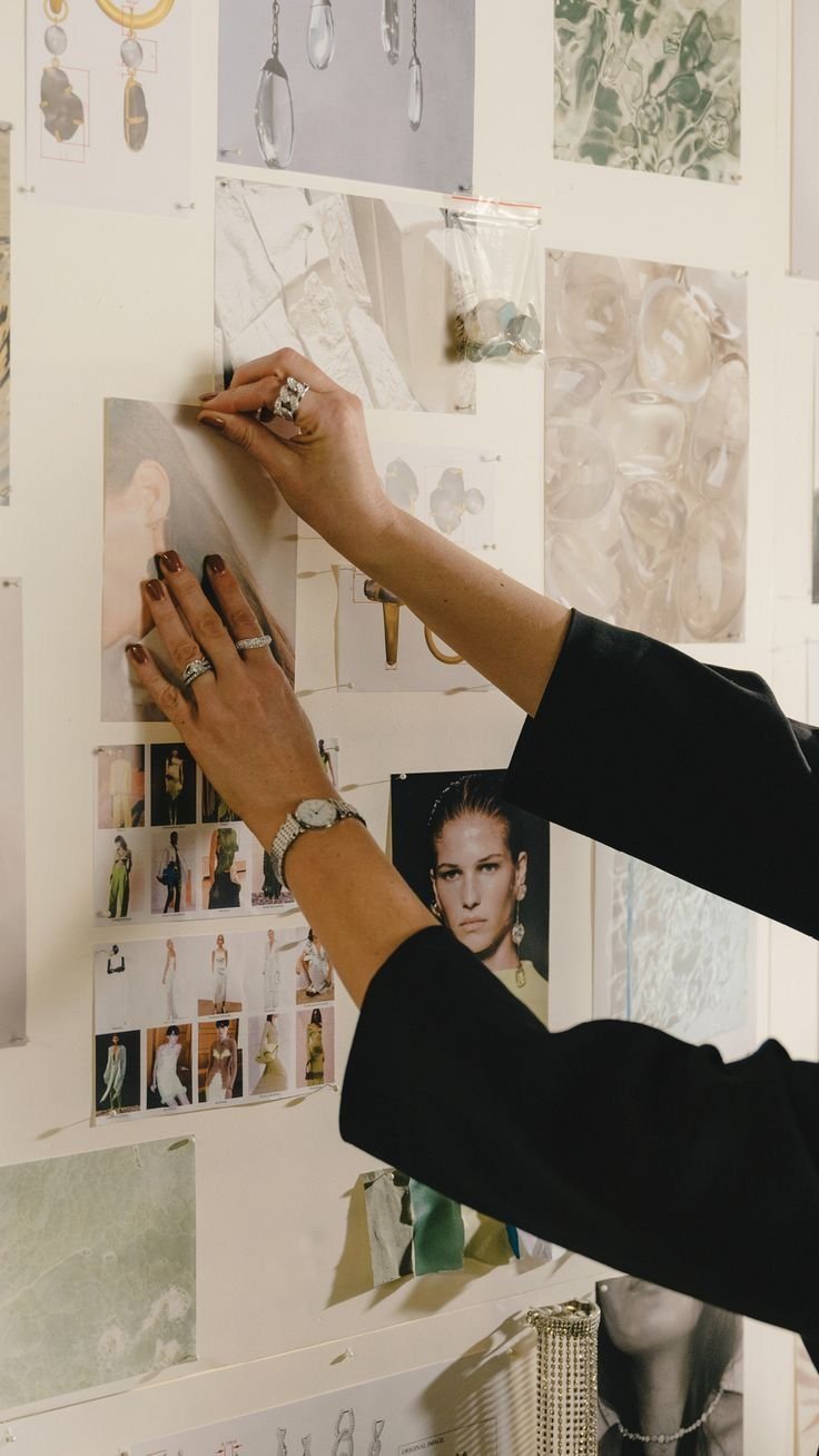 A person is pinning up or arranging photos and images on a wall, including fashion photos, jewelry, and artwork, while wearing rings and a watch.