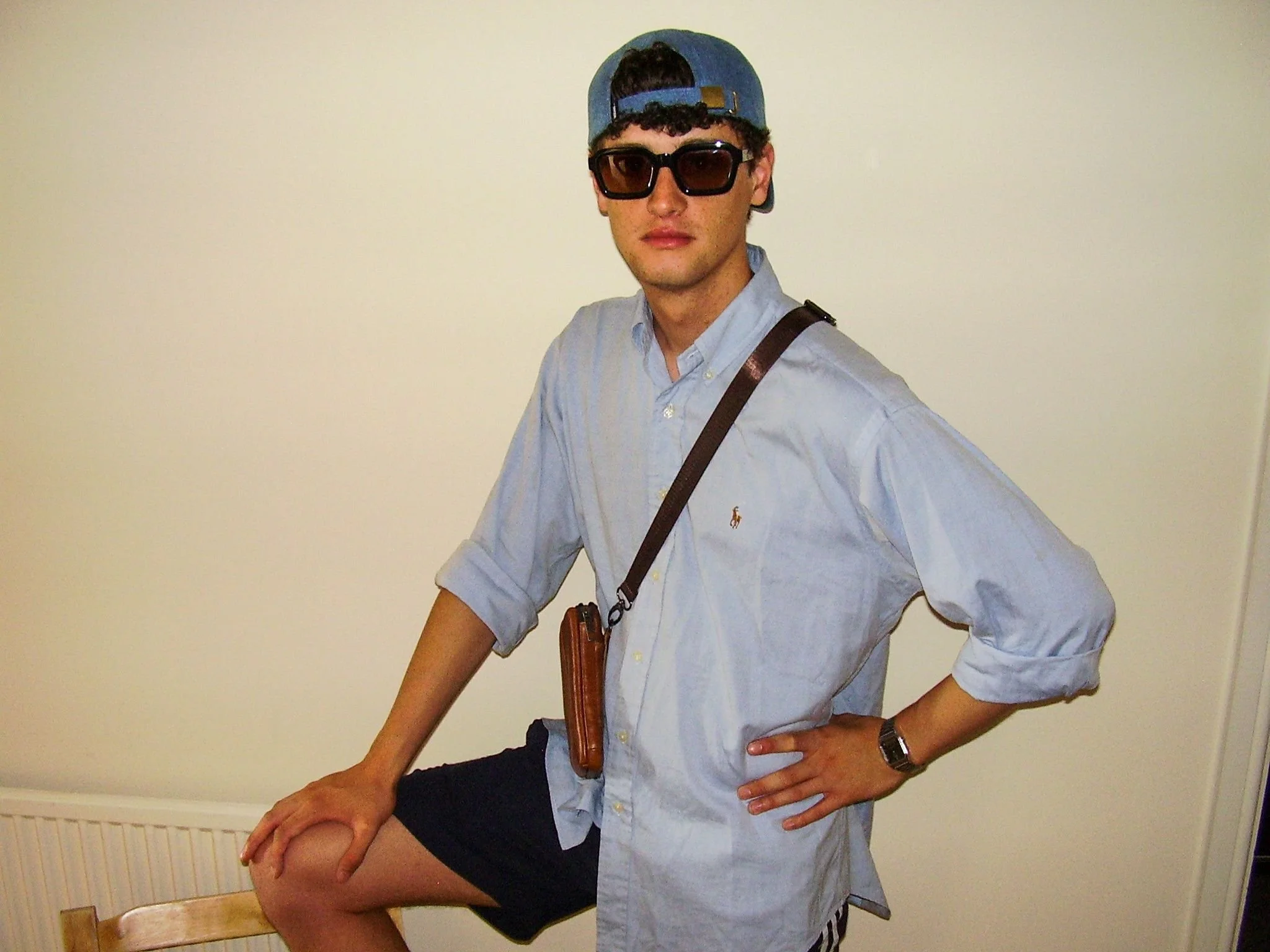 Young man wearing sunglasses, a blue baseball cap backward, a light blue button-down shirt, black shorts, and a wristwatch, standing with one hand on his hip and his other leg resting on a chair.