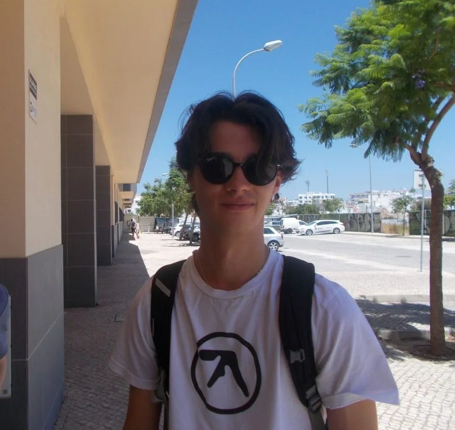 Young man with dark sunglasses, white t-shirt with black logo, carrying a black backpack, standing outdoors on a sunny day near trees and parked cars.