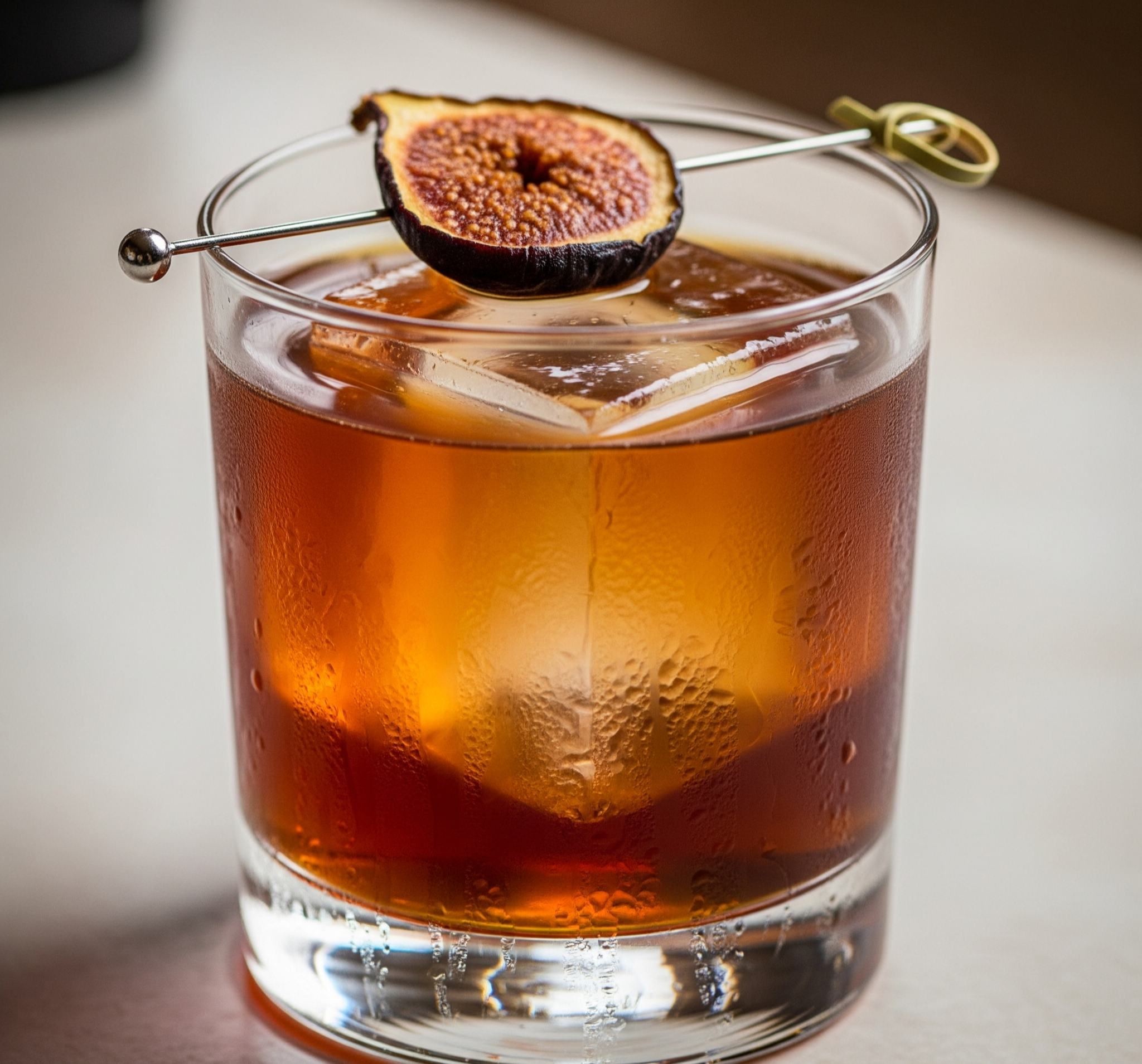 A glass of iced dark amber cocktail with a dried fig garnish on a cocktail pick.