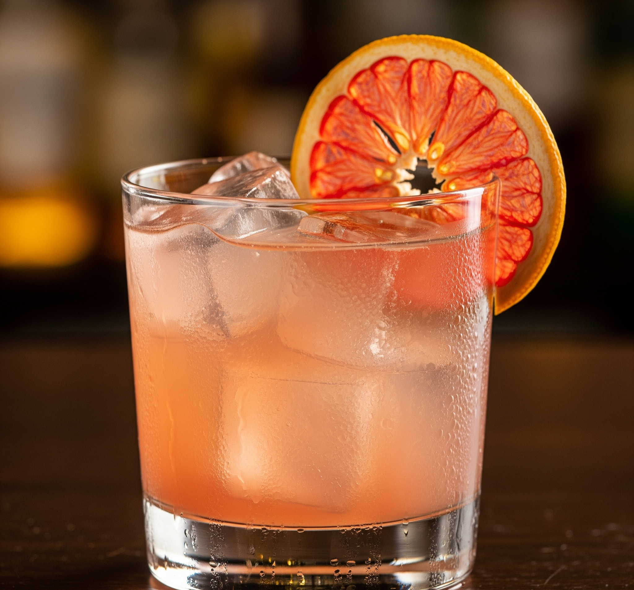 A pink-colored cocktail with ice cubes in a clear glass, garnished with a dried orange slice.