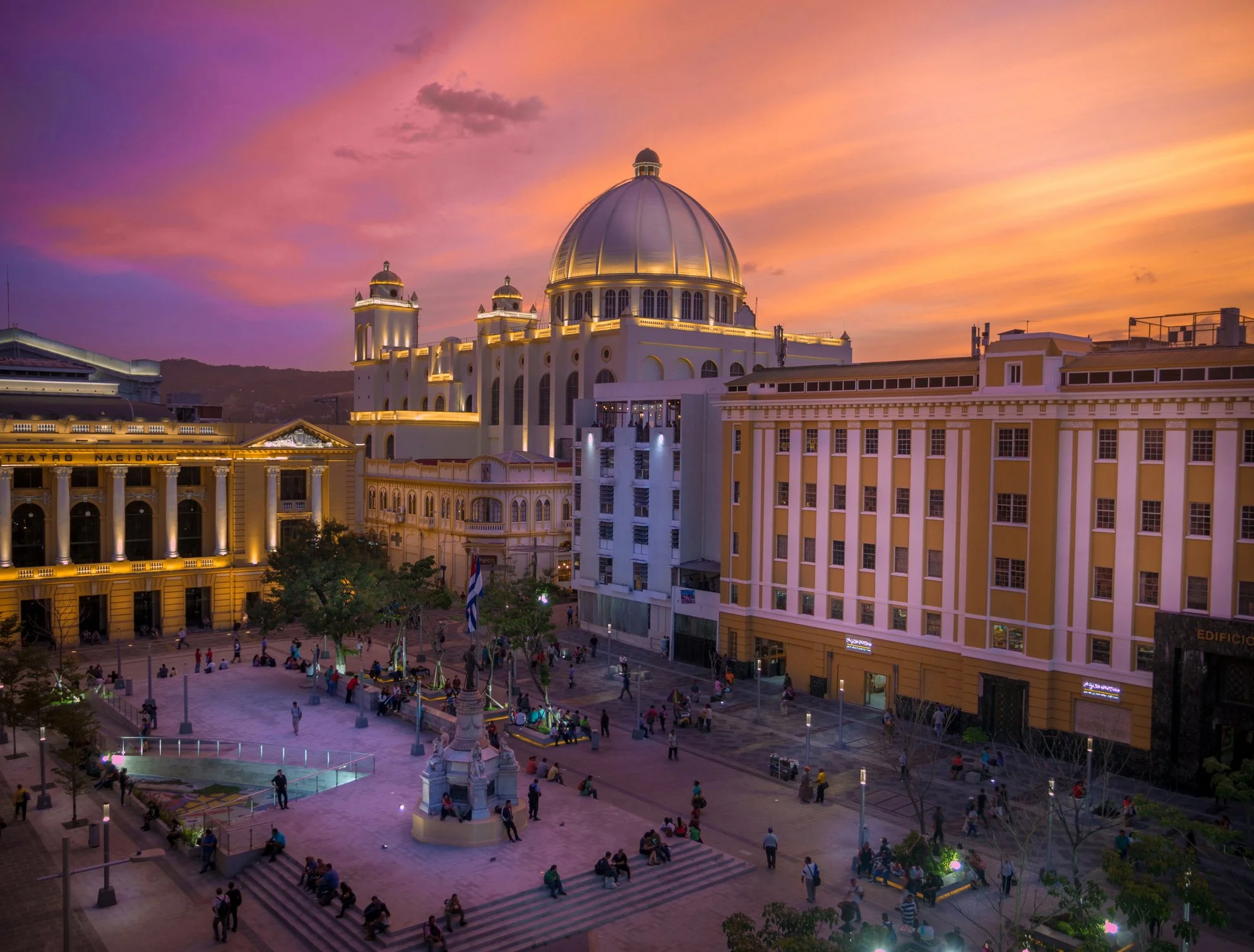 City square at sunset with a large domed building and yellow/white architecture, people sitting and walking, trees, flags, with pink and orange sky.