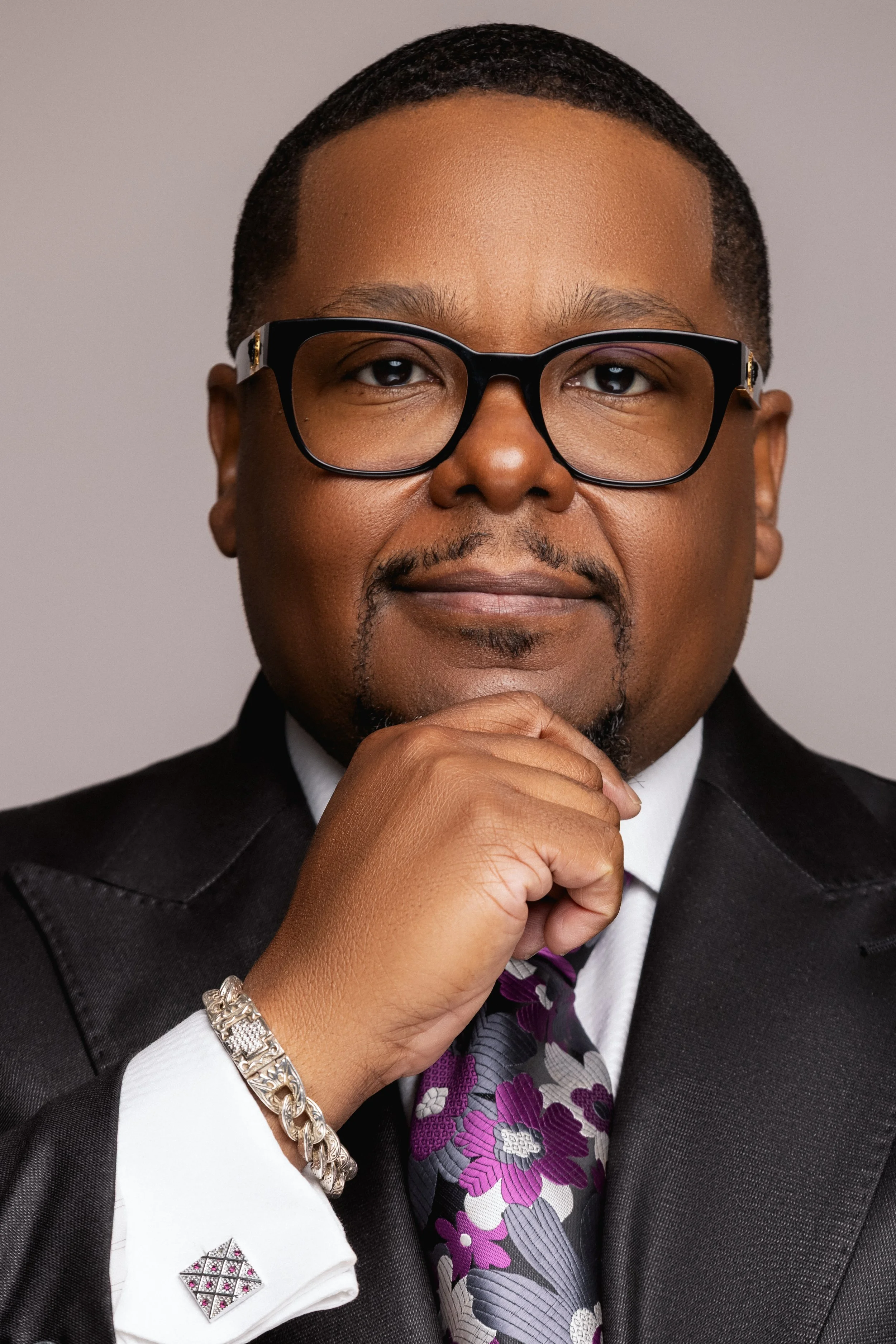 Close-up of a man wearing glasses, a suit, and a purple floral tie, with a thoughtful expression, resting his chin on his hand, against a neutral background.