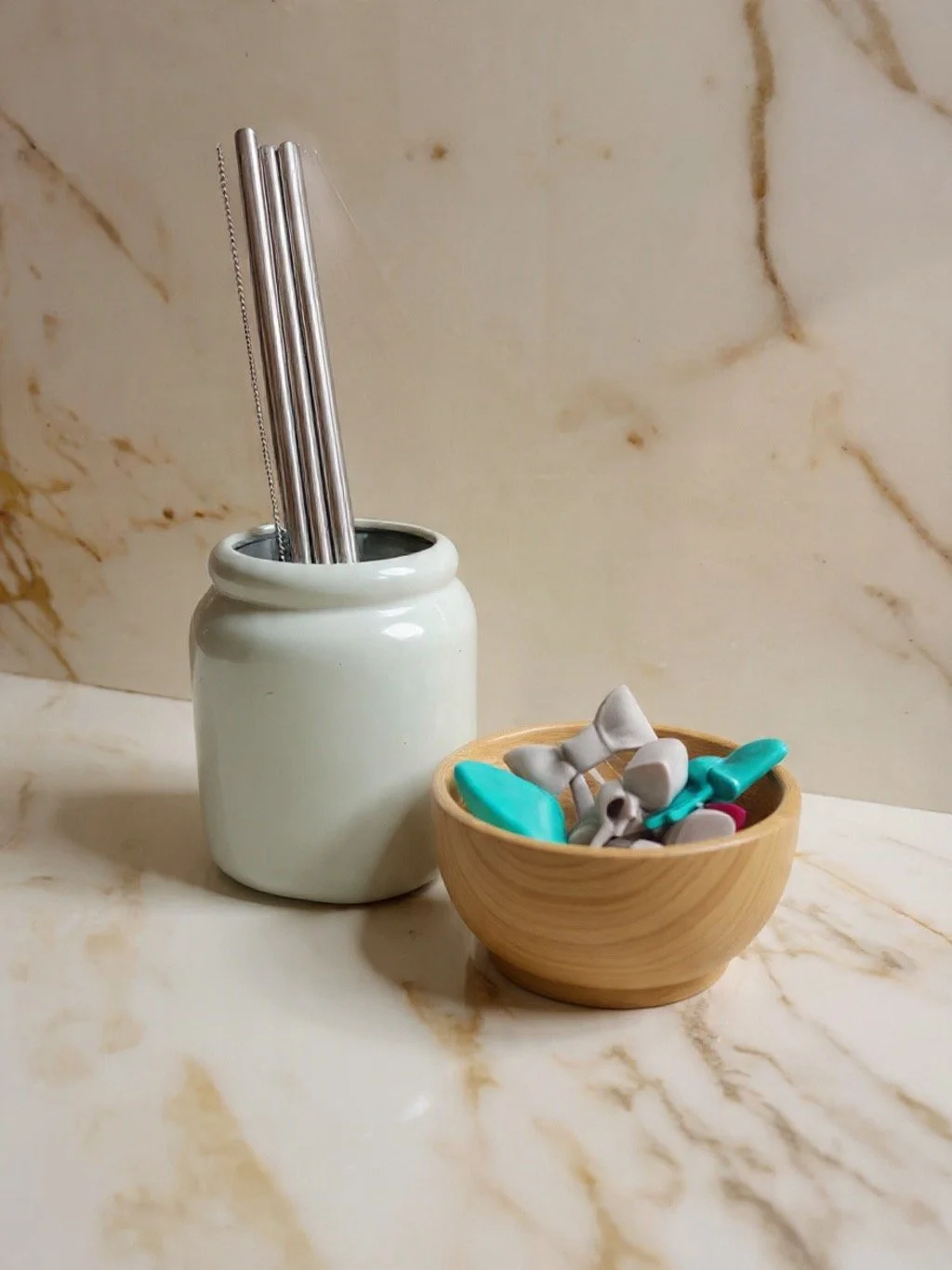 a wood bowl filled with gray and teal bow and whale tail straw attachments on a marble counter adjacent to a teal ceramic cup filled with stainless steel straw attachments