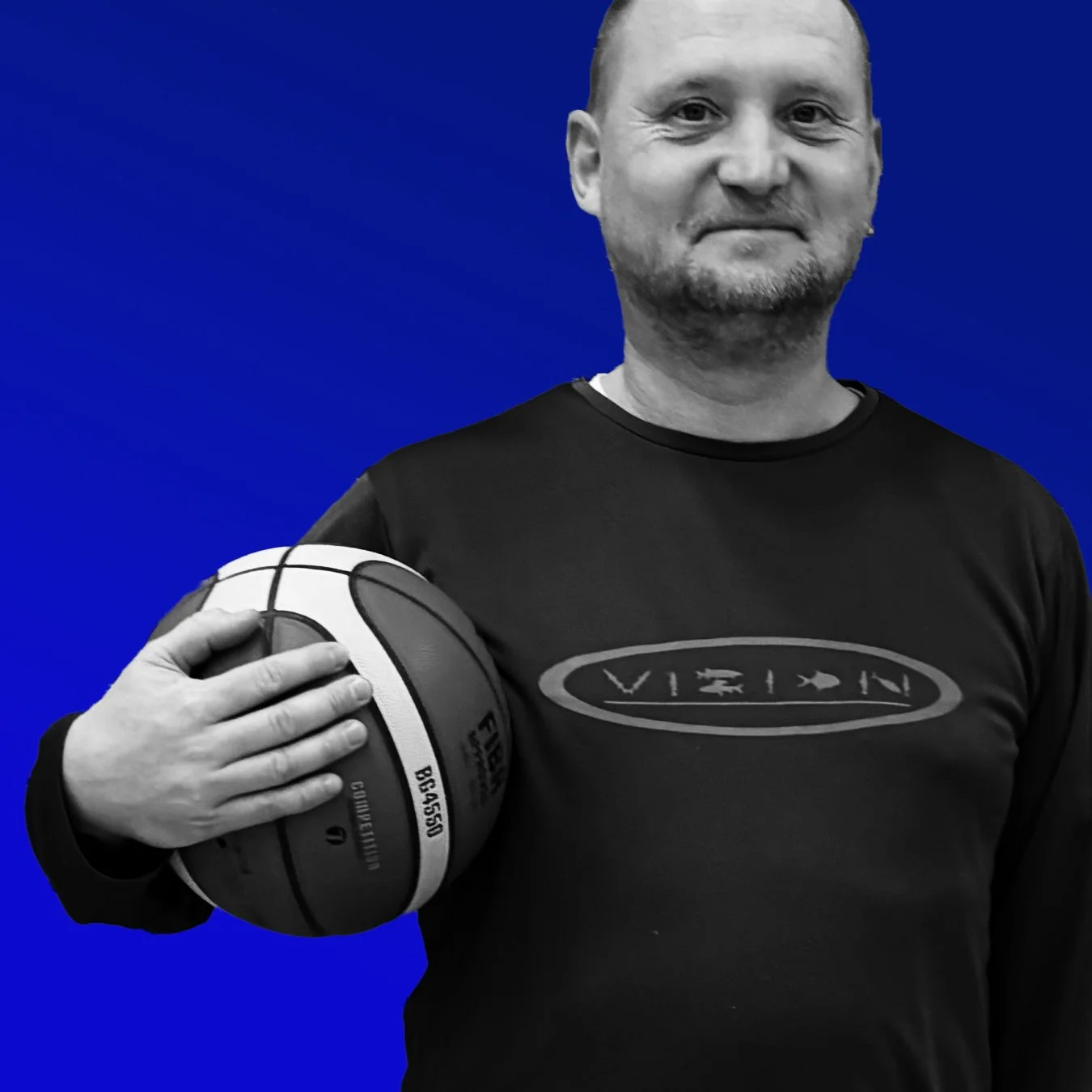 Ein Mann hält einen schwarzen und weißen Basketball mit der linken Hand vor einer blauen Hintergrundwand. Er trägt ein schwarzes T-Shirt mit einer ovalen Marke in der Mitte.