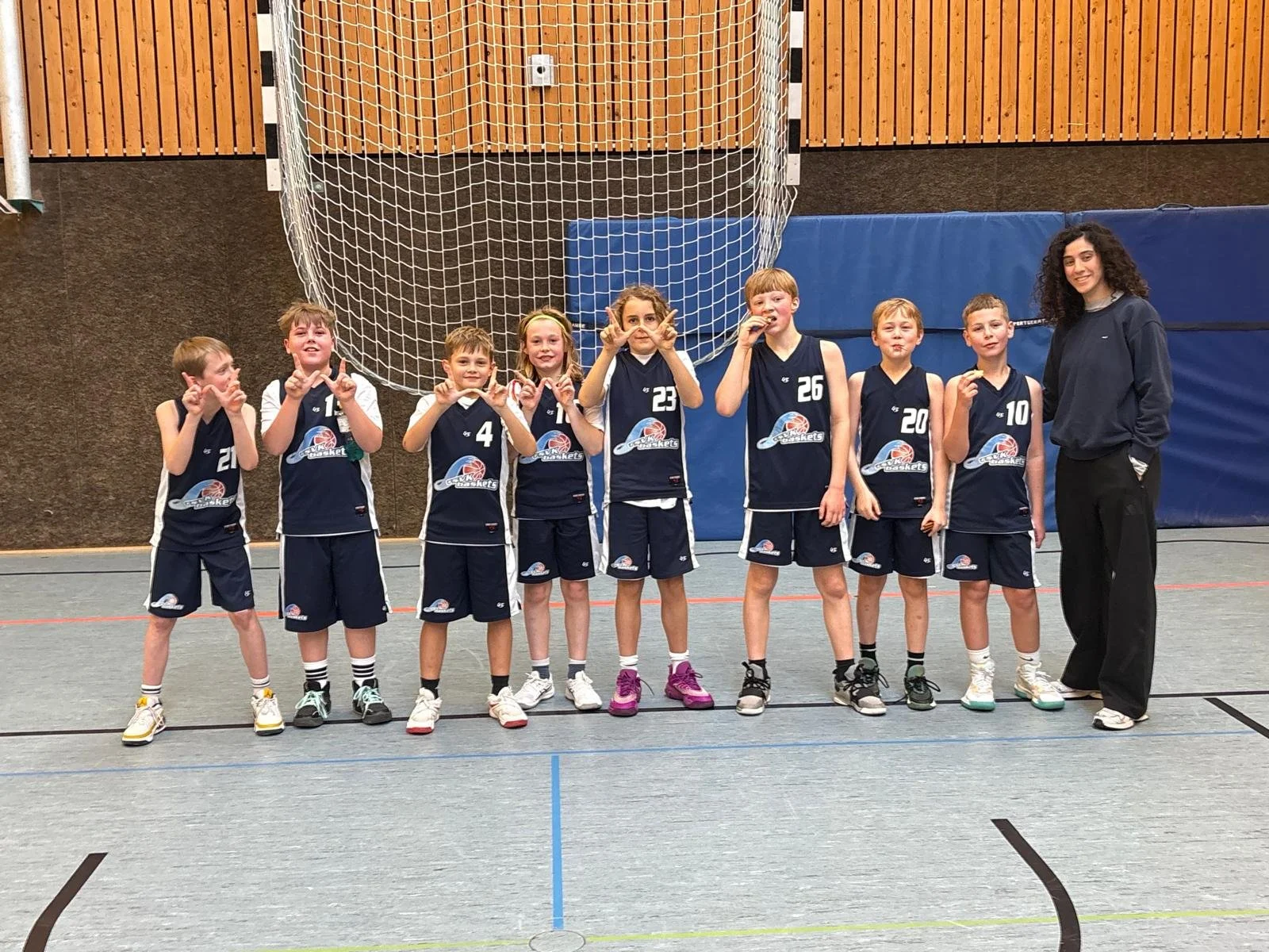 Jugendbasketballteam mit Trainerin in einem Indoor-Sporthalle fotografiert, alle tragen dunkelblaue Trikots mit Nummern und einem Basketball-Logo, die Spieler posieren für das Gruppenfoto.