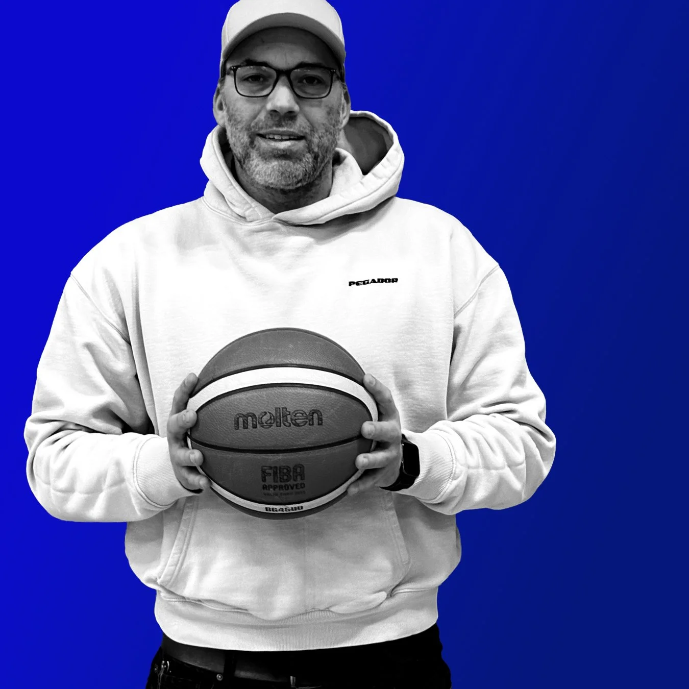 Mann mit Brille, Bart und weißen Kapuzenpullover, hält einen Basketball vor einem blauen Hintergrund.