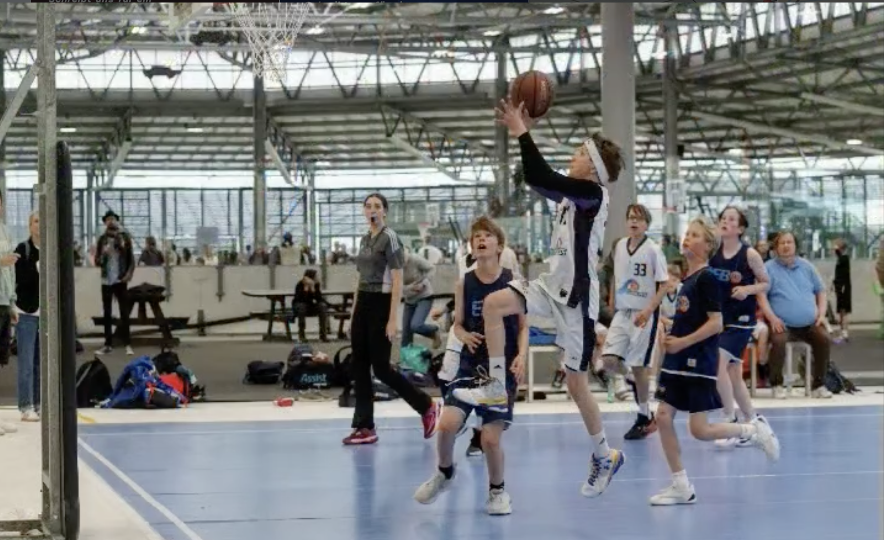 Kinder spielen Basketball in einer Sporthalle, während im Hintergrund Zuschauer sitzen und stehen.