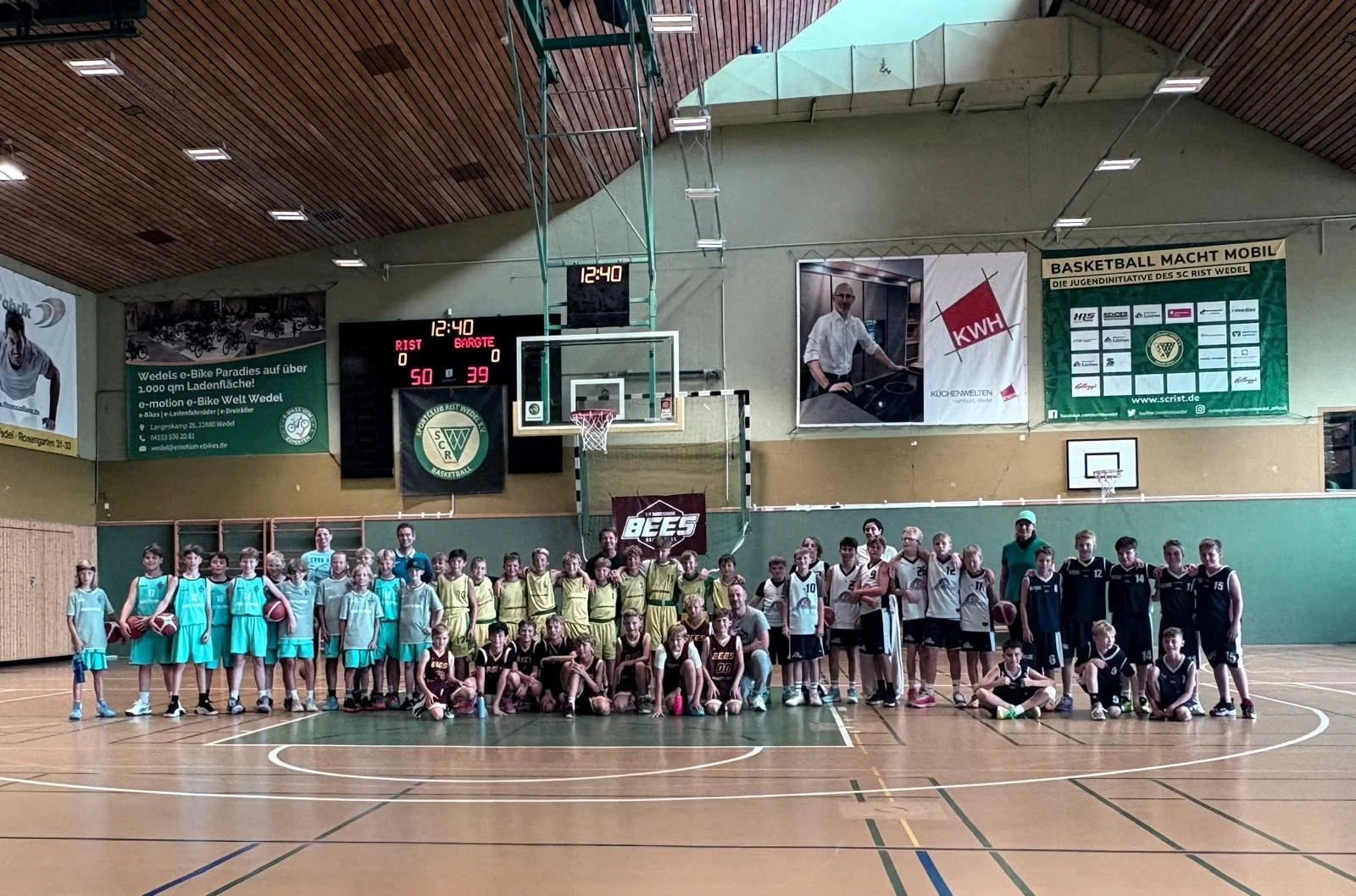 Gruppens Foto von Jugendlichen und Trainern in einer Basketballhalle nach einem Spiel, mit Spielern in blauen, gelben und schwarzen Trikots sowie einem Basketballkorb im Hintergrund.