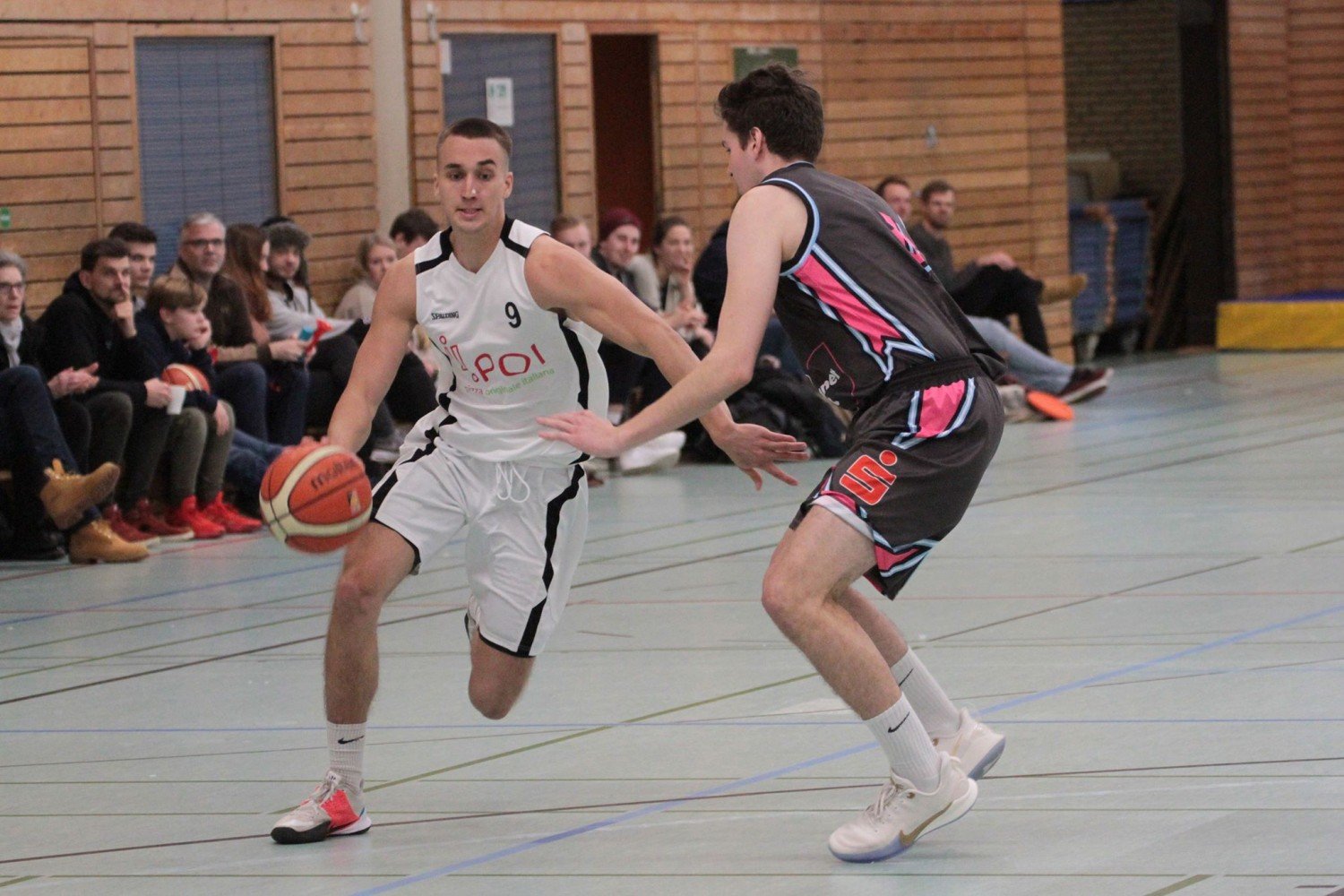 Zwei Basketballspieler im Kampf um den Ball während eines Spiels in einer Turnhalle, im Hintergrund Zuschauer.