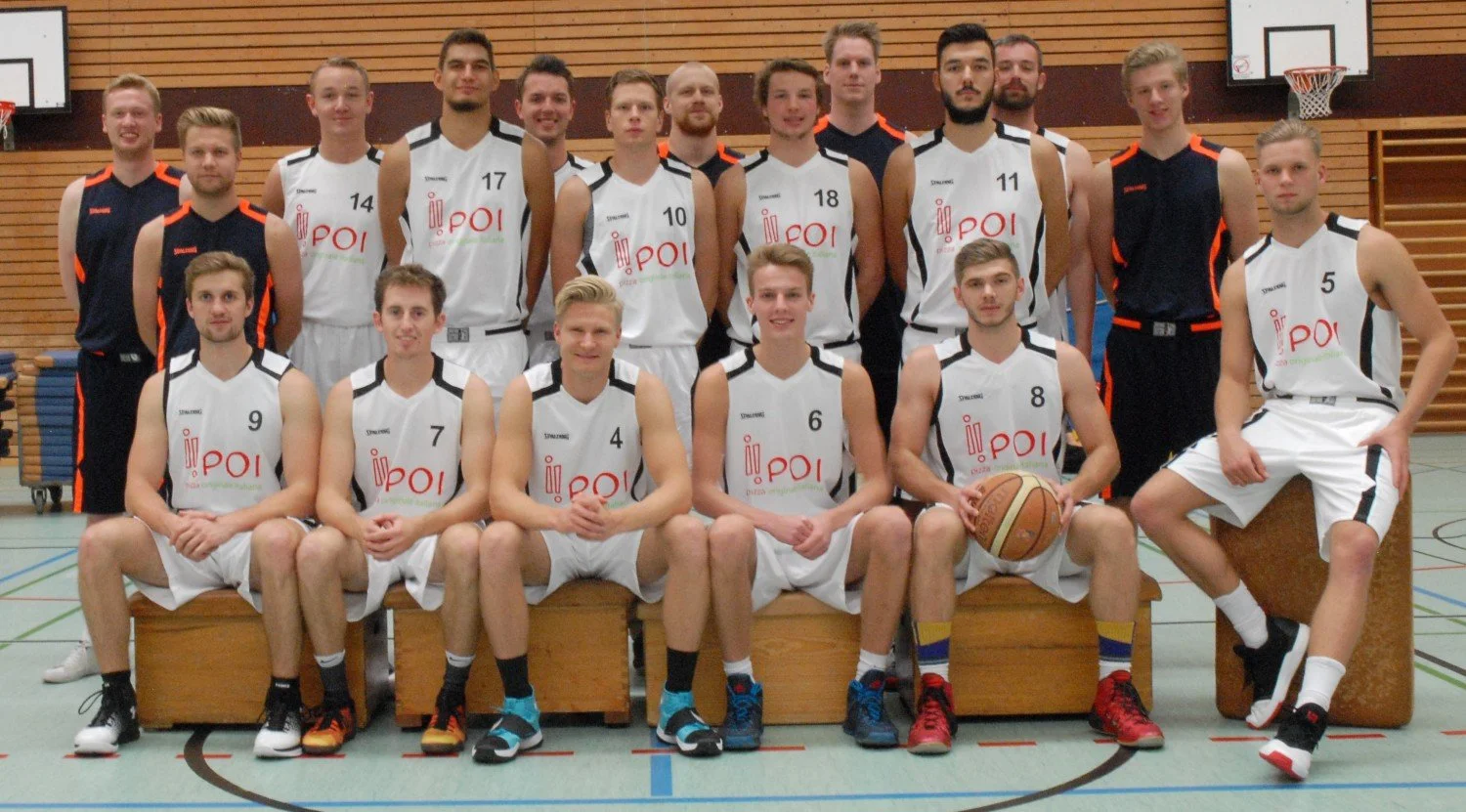 Basketballmannschaft in Sporttrikots im Innenraum der Turnhalle, einige sitzen, andere stehen, mit Basketball und Turnbänken.