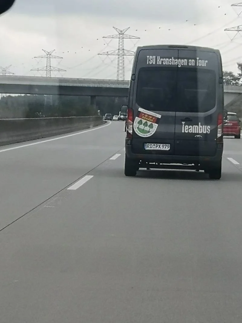 Ein schwarzer Van fährt auf der Autobahn. Auf der Rückseite des Vans steht "150 Kreuzhagen on Tour" und "Teambus". Es gibt auch ein Schild mit einem Rote-Punkt-Logo und einer grünen Baumgruppe.