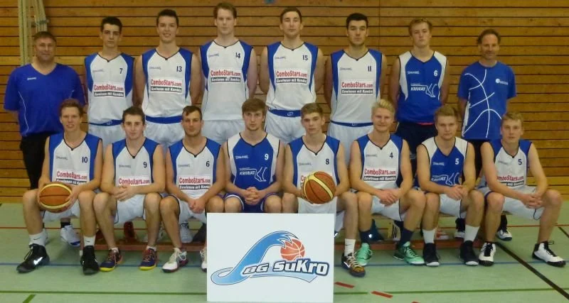 Eine Basketballmannschaft mit 13 Spielern und 2 Trainern posiert in einer Turnhalle. Die Spieler tragen blaue und weiße Trikots mit Nummern, einige halten Basketbälle.