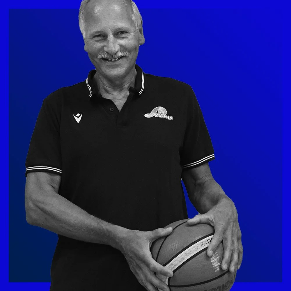 Älterer Mann mit grauem Haar und Bart, der einen Basketball hält, vor blauem Hintergrund. Er trägt ein schwarzes Sportshirt mit Logo.