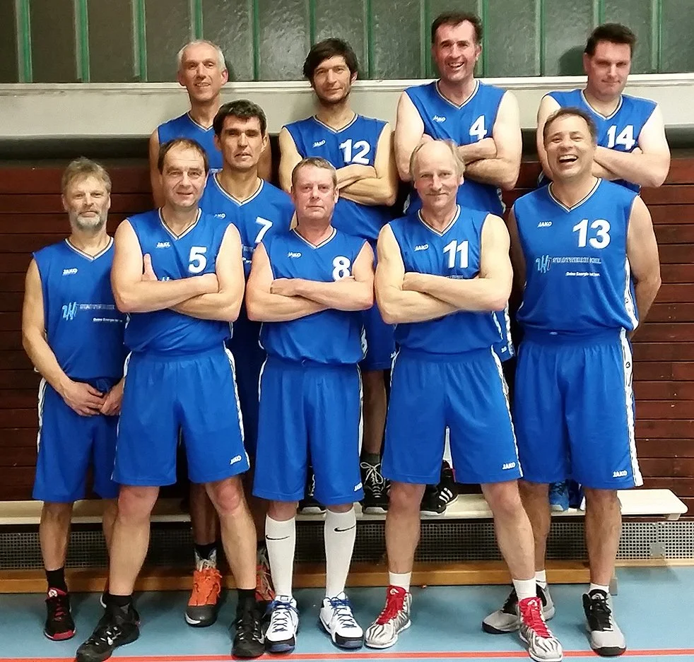 Eine Gruppe von männlichen Volleyballspielern in blauen Trikots steht in einer Halle vor einer Holzwand, einige haben die Arme verschränkt, alle tragen Sportschuhe.