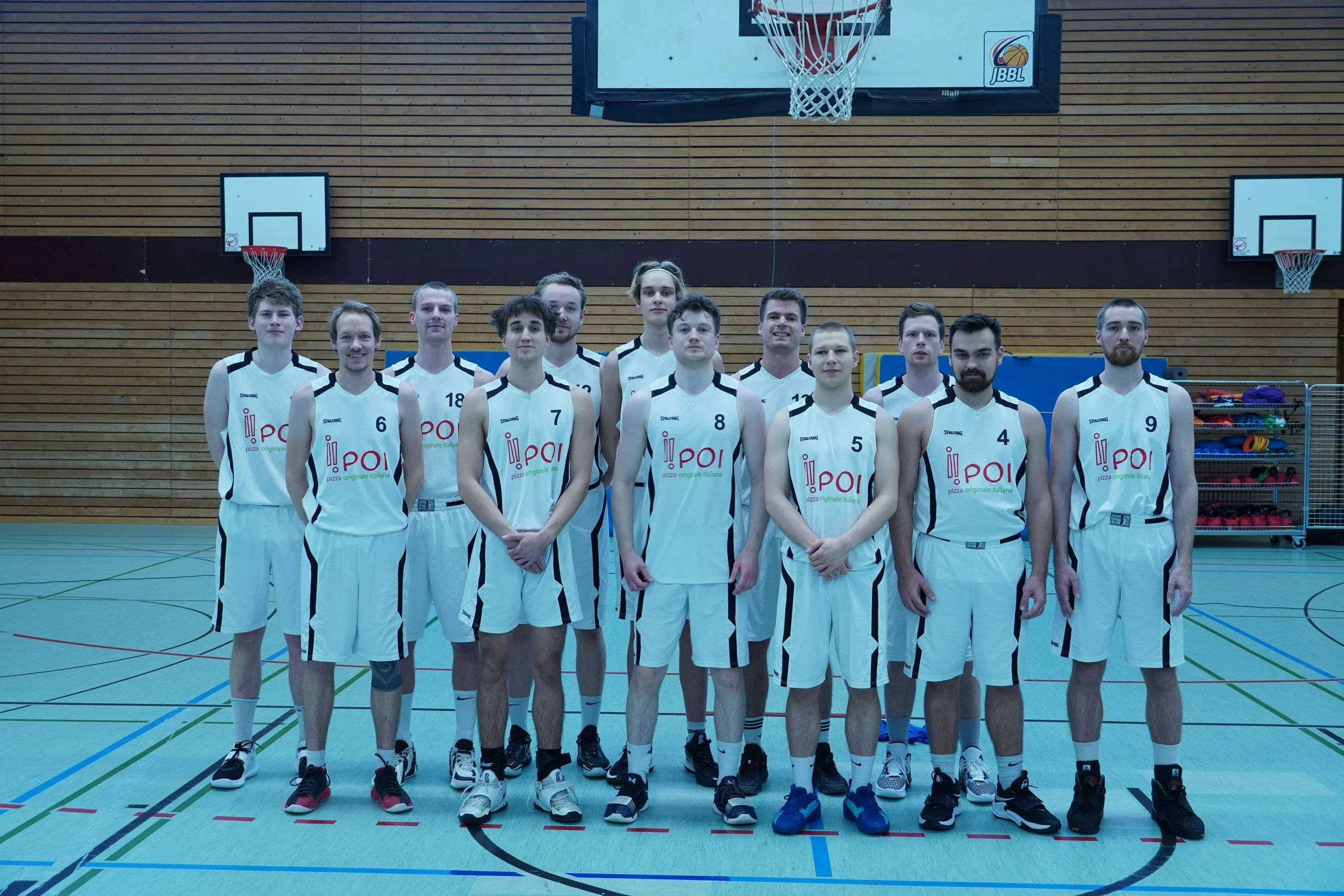 Ein Basketballteam posiert in einer Turnhalle vor einem Basketballkorb für ein Foto. Die Spieler tragen weiße Trikots mit schwarzen Details.