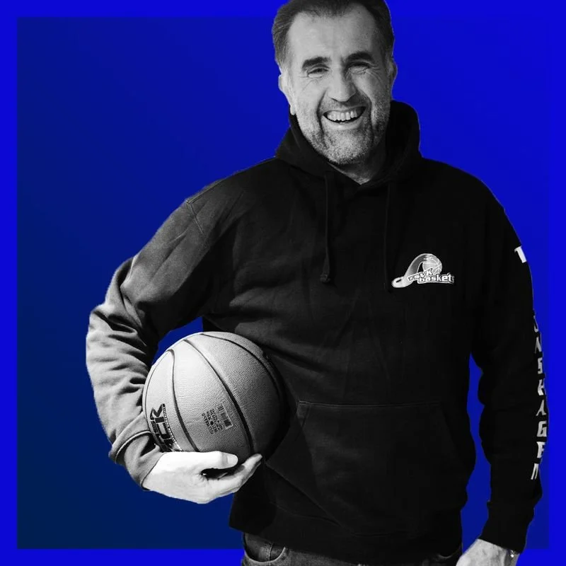Mann in schwarzem Hoodie, lächelnd, hält Basketball vor einem blauen Hintergrund.