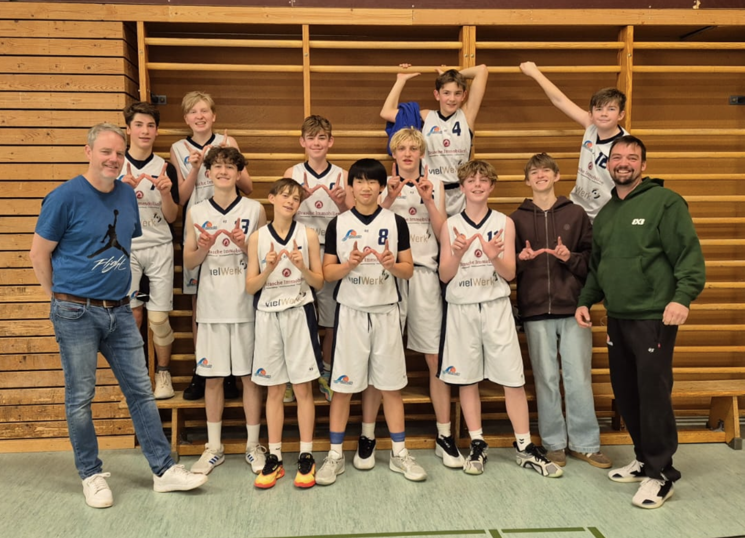 Jugendbasketballteam mit zwei Erwachsenen, alle in weißen Trikots, stehen vor einer Holztrainingstafel in der Turnhalle, some team members sind in der vorderen Reihe, einige sind auf einer höheren Ebene im Hintergrund, alle lächeln und machen Herzhan