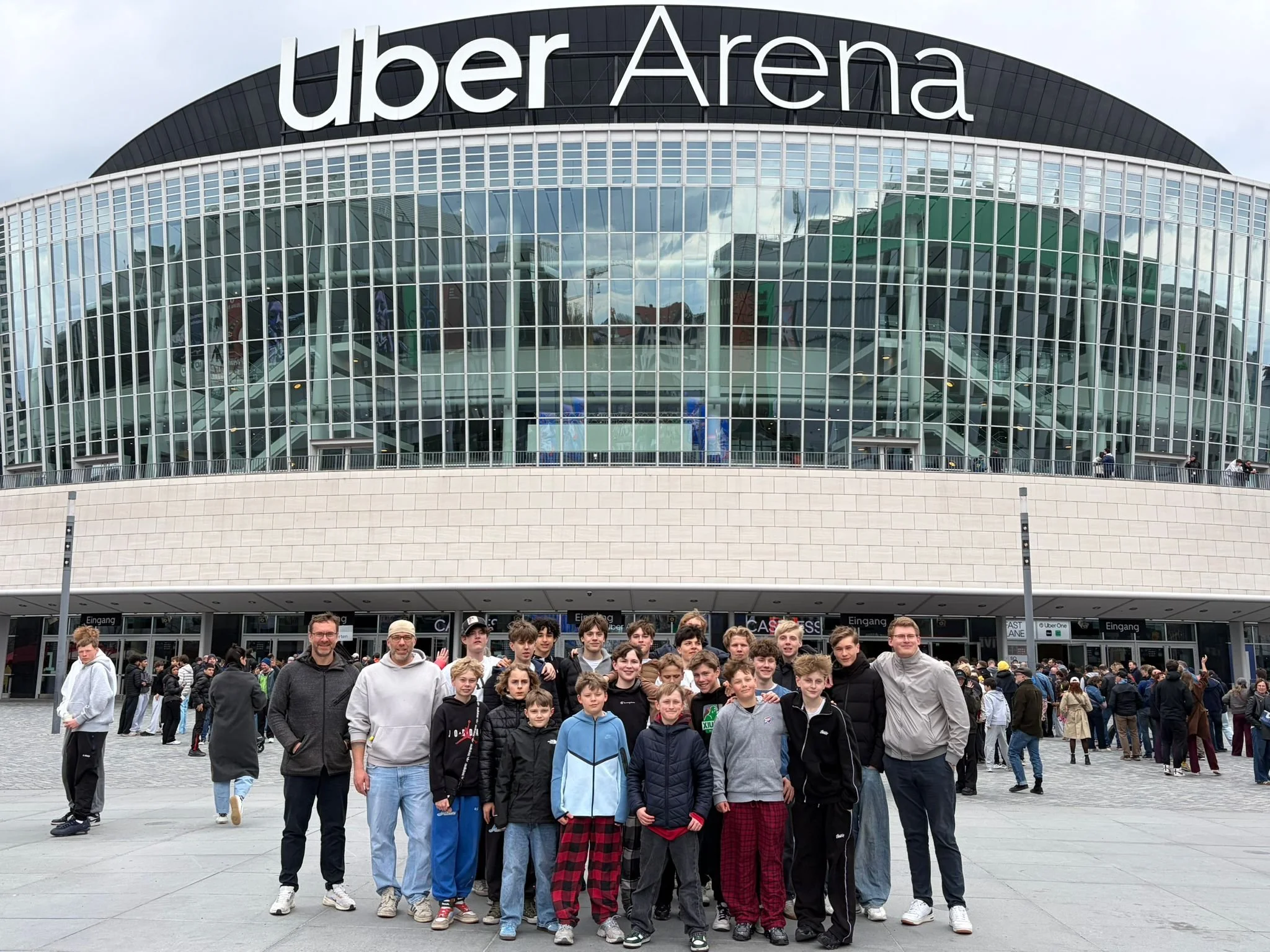 Eine Gruppe junger Menschen und Erwachsene vor dem Eingang des Berliner Flughafens, genannt "über Arena", mit einem modernen Glasgebäude im Hintergrund.