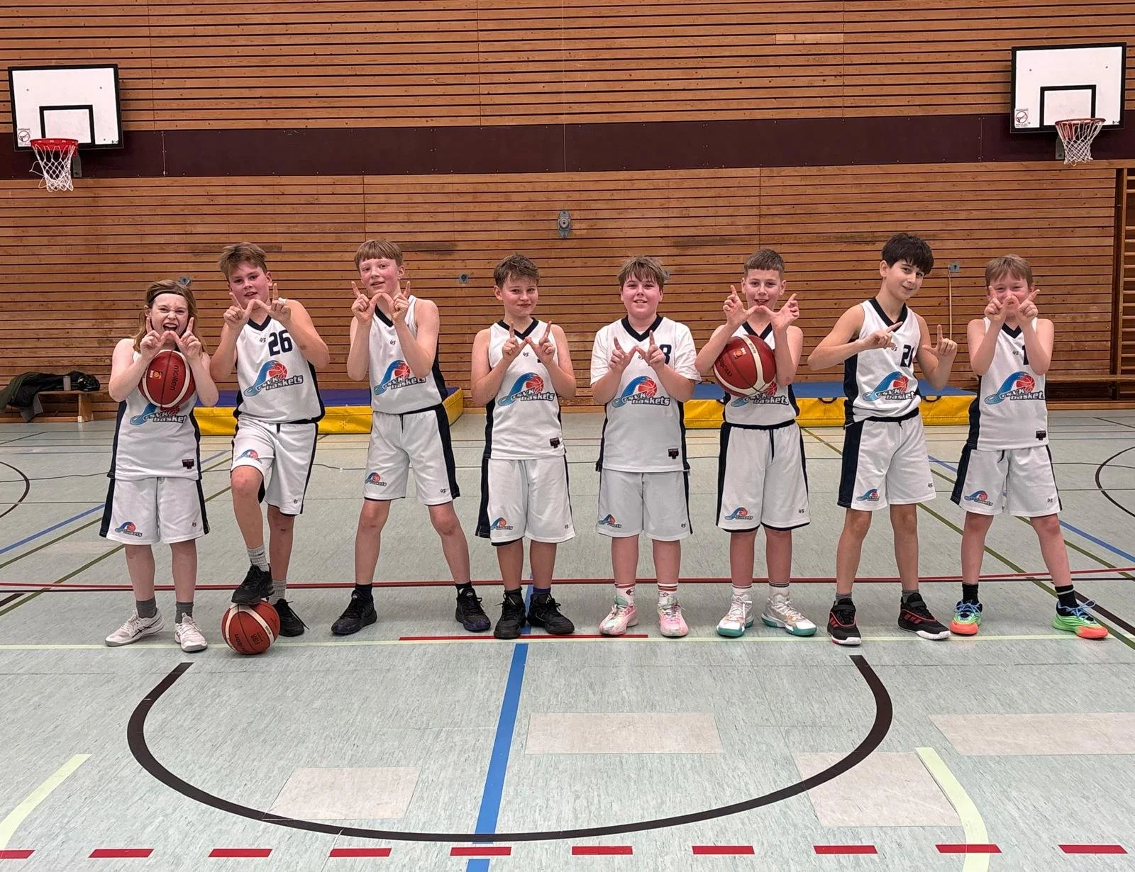 Eine junge Basketballmannschaft besteht aus acht Kindern in weißen Trikots und Shorts, die in einer Turnhalle stehen. Zwei Kinder halten Basketballs, während die anderen die Hände in verschiedenen Gesten zeigen. Die Kinder lächeln und posieren für da
