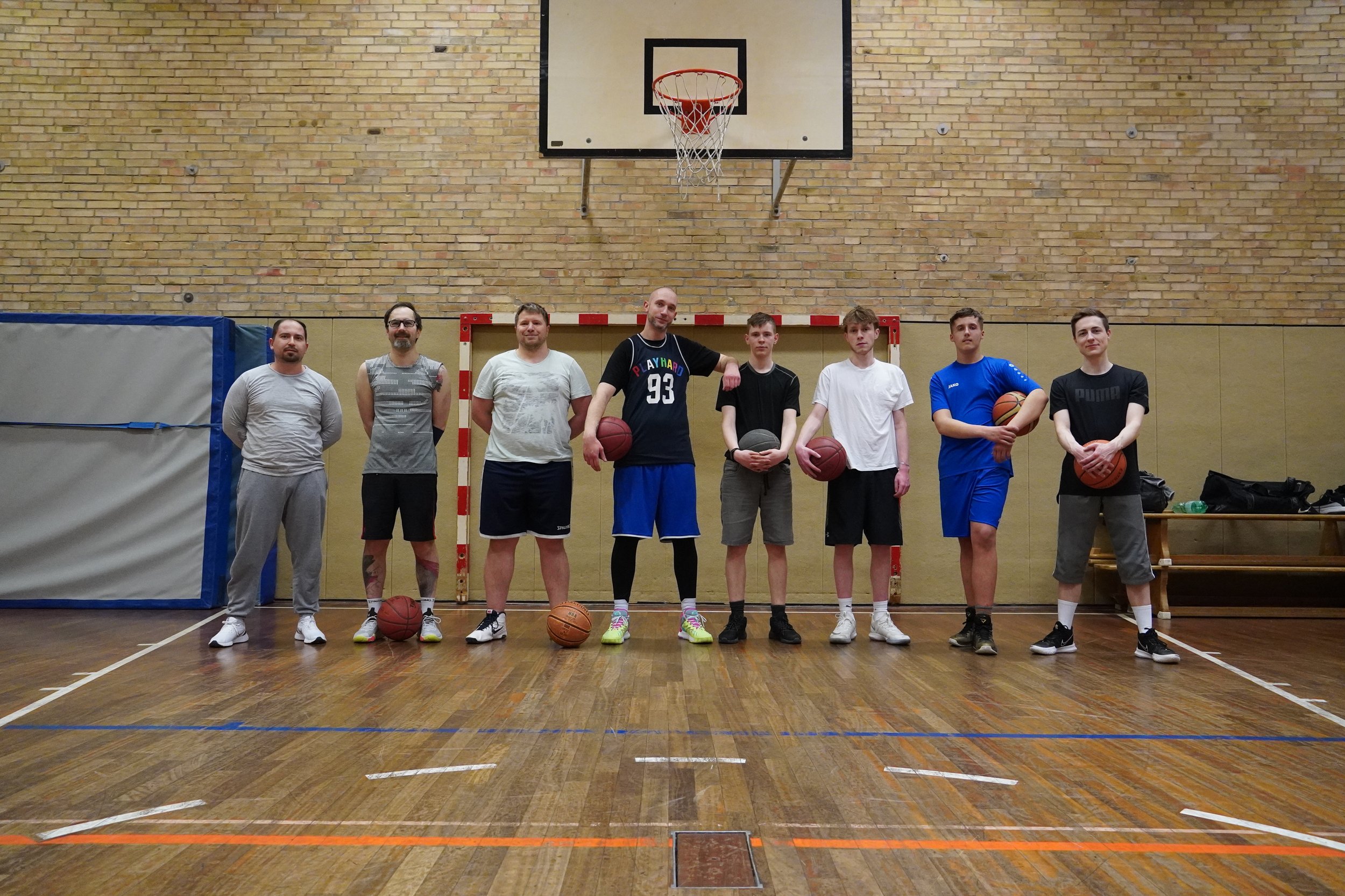 Sieben Männer stehen in einer Turnhalle vor einem Basketballkorb. Sie halten Basketballs, tragen Sportkleidung und sind bereit zum Spielen.