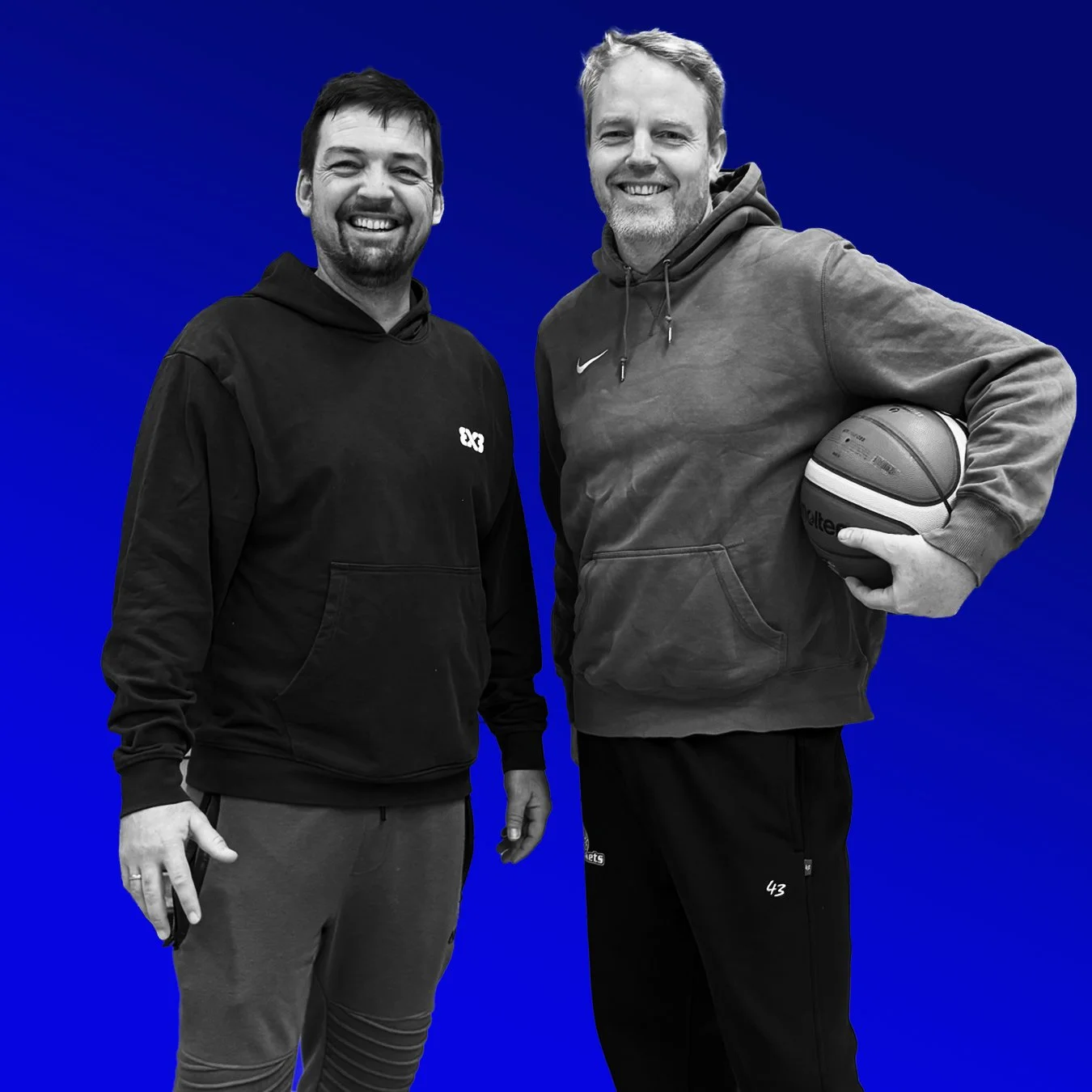 Zwei lachende Männer stehen nebeneinander vor einem blauen Hintergrund. Der Mann auf der rechten Seite hält einen Basketball in der Hand. Beide tragen Kapuzenpullover und Sporthose.