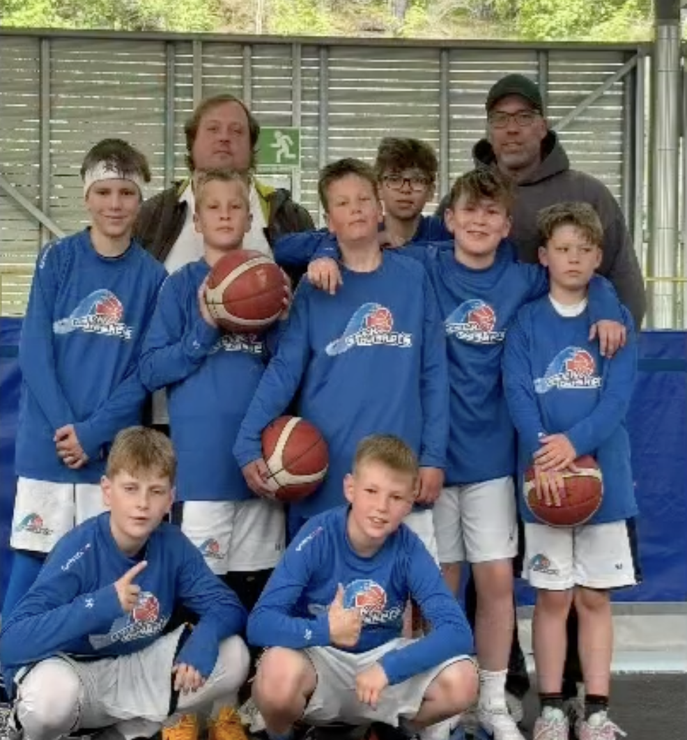 Jugendbasketballteam mit Trainern, alle in blauen Trikots, in einer Halle, einige mit Basketbällen, feiern ihren Erfolg.