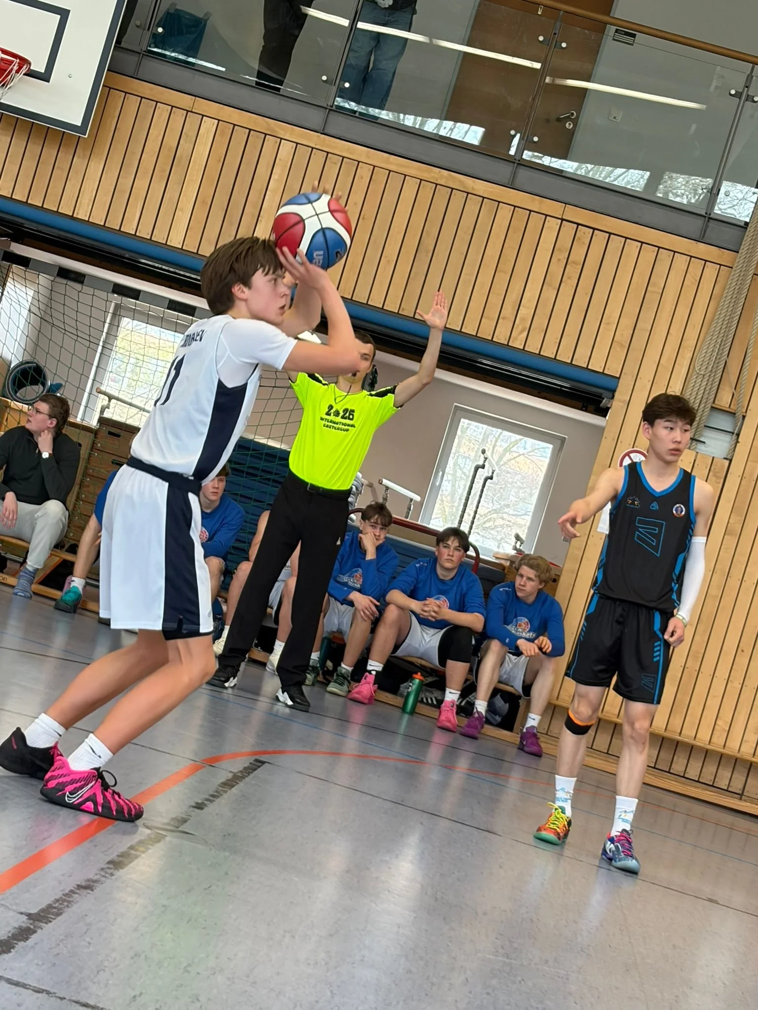 Jugendlicher Spieler beim Volleyballspiel im Innenraum. Er wirft den Ball in die Luft, während ein Schiedsrichter im Neon-Gelb-Kleid die Aktion beobachtet. Im Hintergrund sitzen Jugendliche auf einer Bank, mit Blick auf das Spiel.