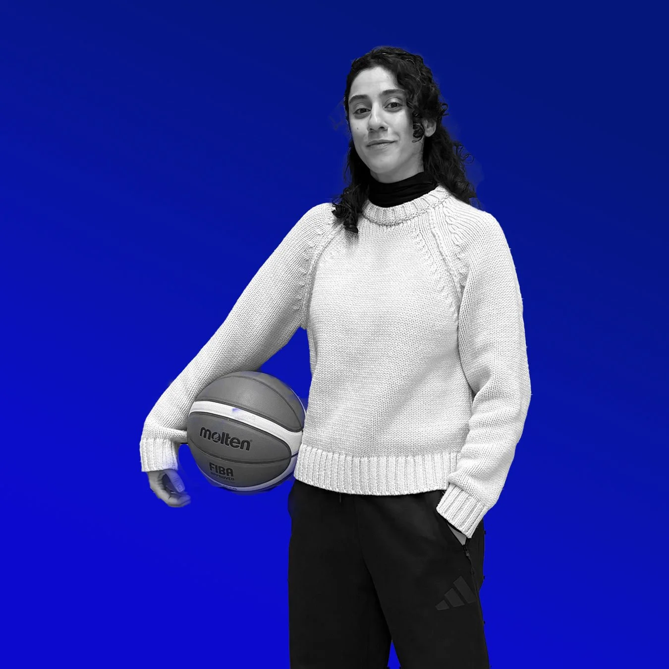 Frau in schwarzer Jogginghose und weißem Pullover hält einen Basketball, steht vor einem blauen Hintergrund.
