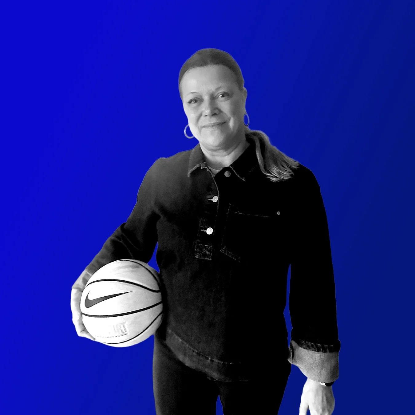 Frau mit kurzem Haar in dunkler Jacke, hält einen Basketball mit Nike-Logo vor einem blauen Hintergrund.