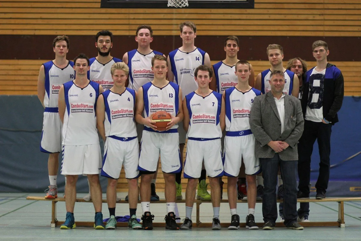 Eine Basketballmannschaft mit roten Trikots und zwei Betreuern steht für ein Gruppenfoto in einer Turnhalle vor einer Holzwand.