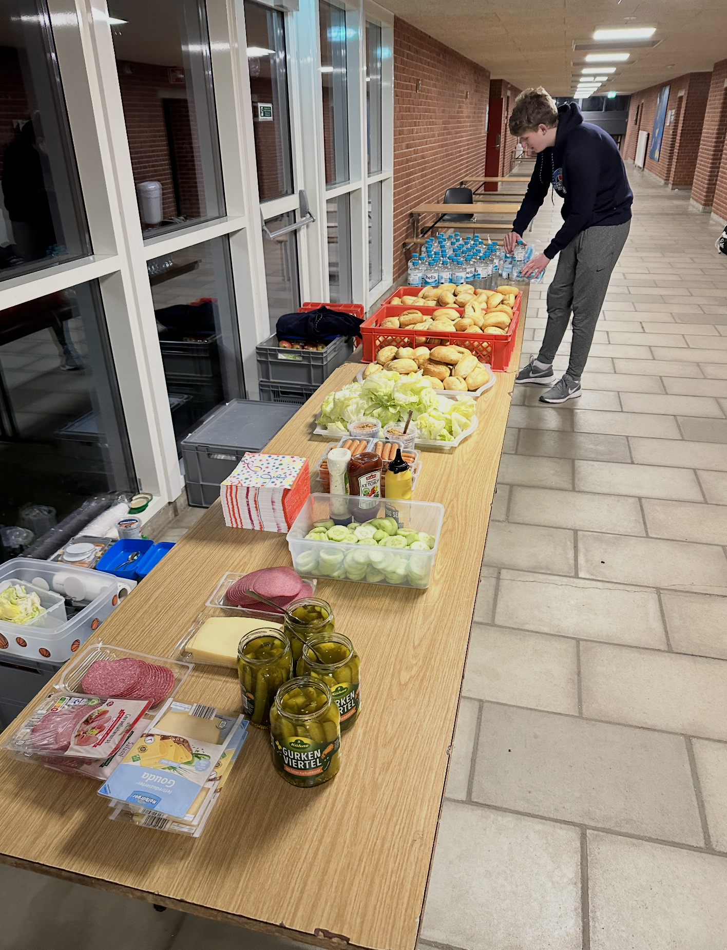 Lebensmittel und Getränke auf einem langen Tisch in einem Flur, darunter eingelegtes Gemüse, frischer Salat, Brot, Wurst, Käse, Senf, Ketchup, Getränke, Wasserflaschen, Gurken, Aufschnitt, Papiertücher und Lebensmittelverpackungen.