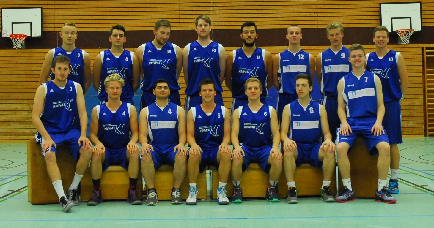 Ein Basketballteam in blauen Trikots posiert für ein Gruppenfoto in einer Turnhalle.
