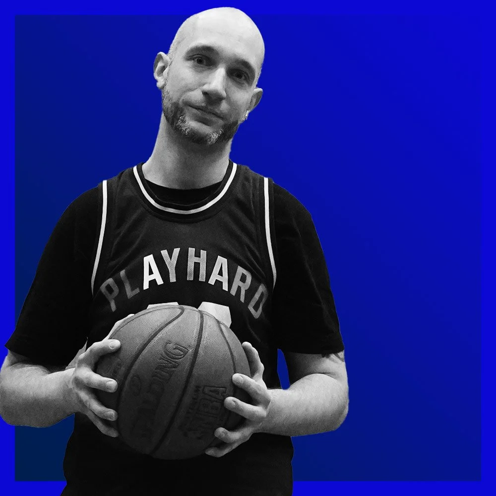 Ein Mann in einem schwarzen Basketballtrikot hält einen Basketball, vor einem blauen Hintergrund.