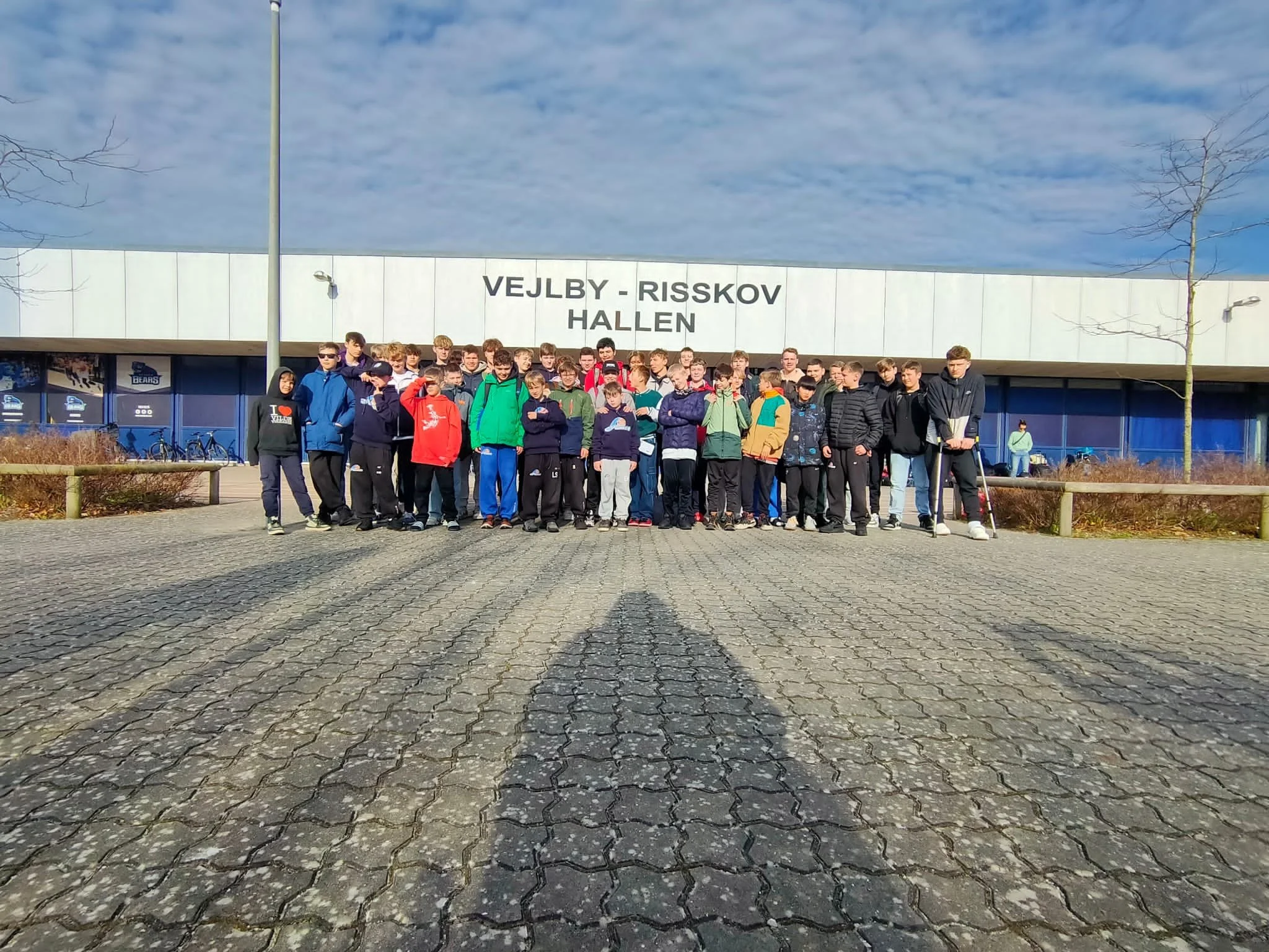 Gruppenjunge vor dem Eingang der Vejlby-Risskov Halle, es ist ein sonniger Tag, keine Menschenmenge sichtbar.