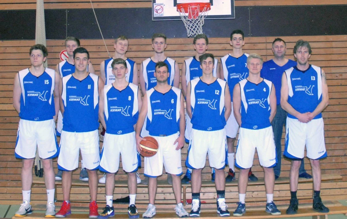 Eine Basketballmannschaft mit zehn Spielern in blauen Trikots und weißen Shorts, steht auf einer Holztribüne vor einem Basketballkorb in einer Sporthalle.