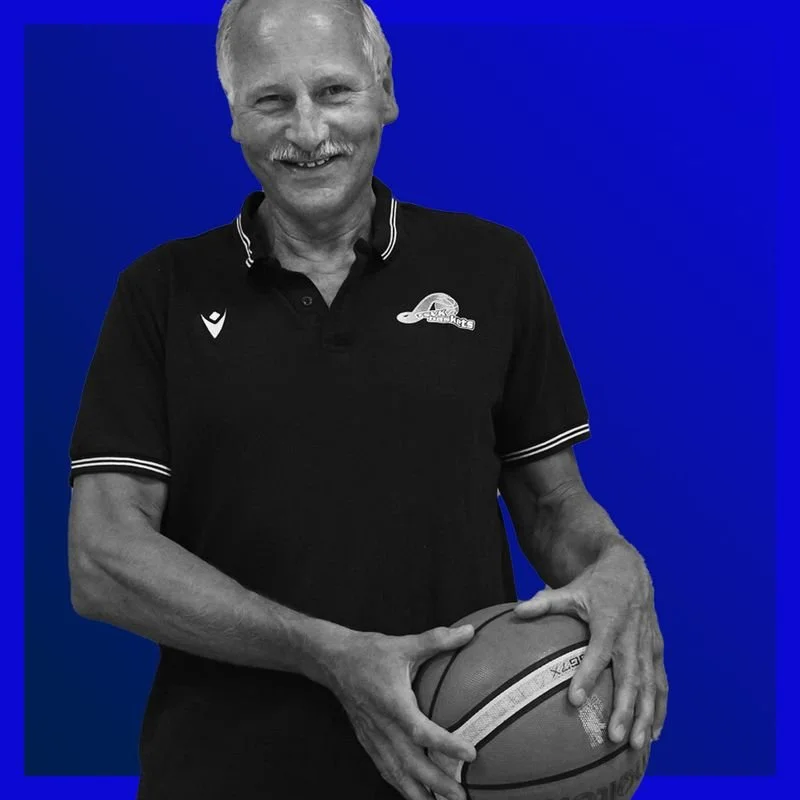 Älterer Mann in schwarzem Polo-Shirt mit Basketball in der Hand, lächelnd vor blauem Hintergrund.