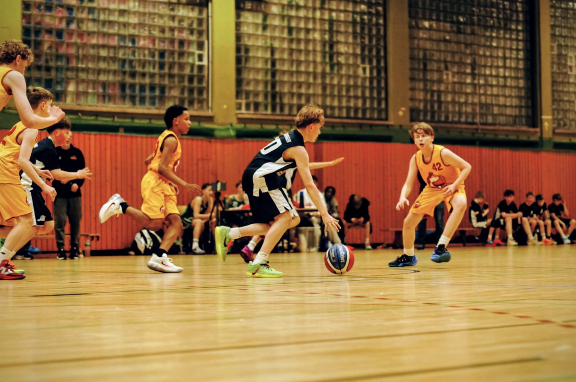 Jugendliche beim Basketballspiel in der Halle, einige in gelben Trikots, einer in schwarz-weiß, einer mit Ball, im Hintergrund Tribünen mit Zuschauern.