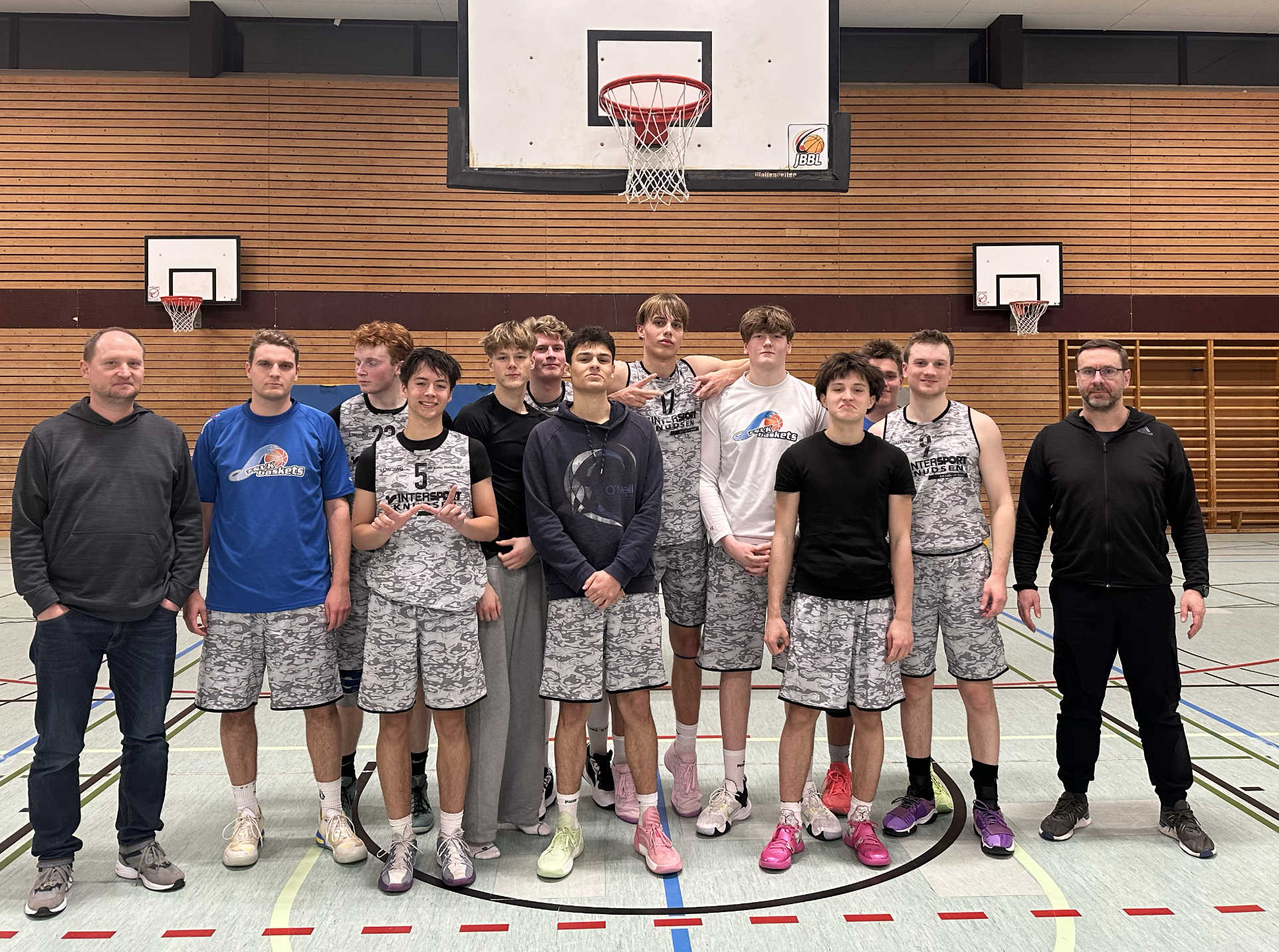 Gruppenfoto einer Basketballmannschaft sowie zwei Trainer in einer Turnhalle, mit Basketballkörben im Hintergrund.
