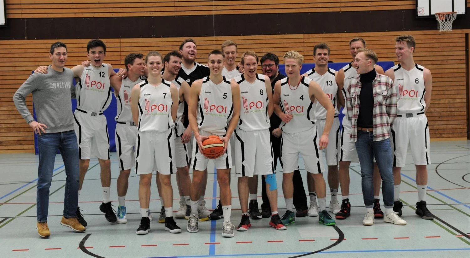 Mannschaftsfoto eines Basketballteams in Weiß mit schwarzen Details, die zusammen in einer Turnhalle stehen, einige lachen, eine Person hält einen Basketball, alle sind sportlich gekleidet.